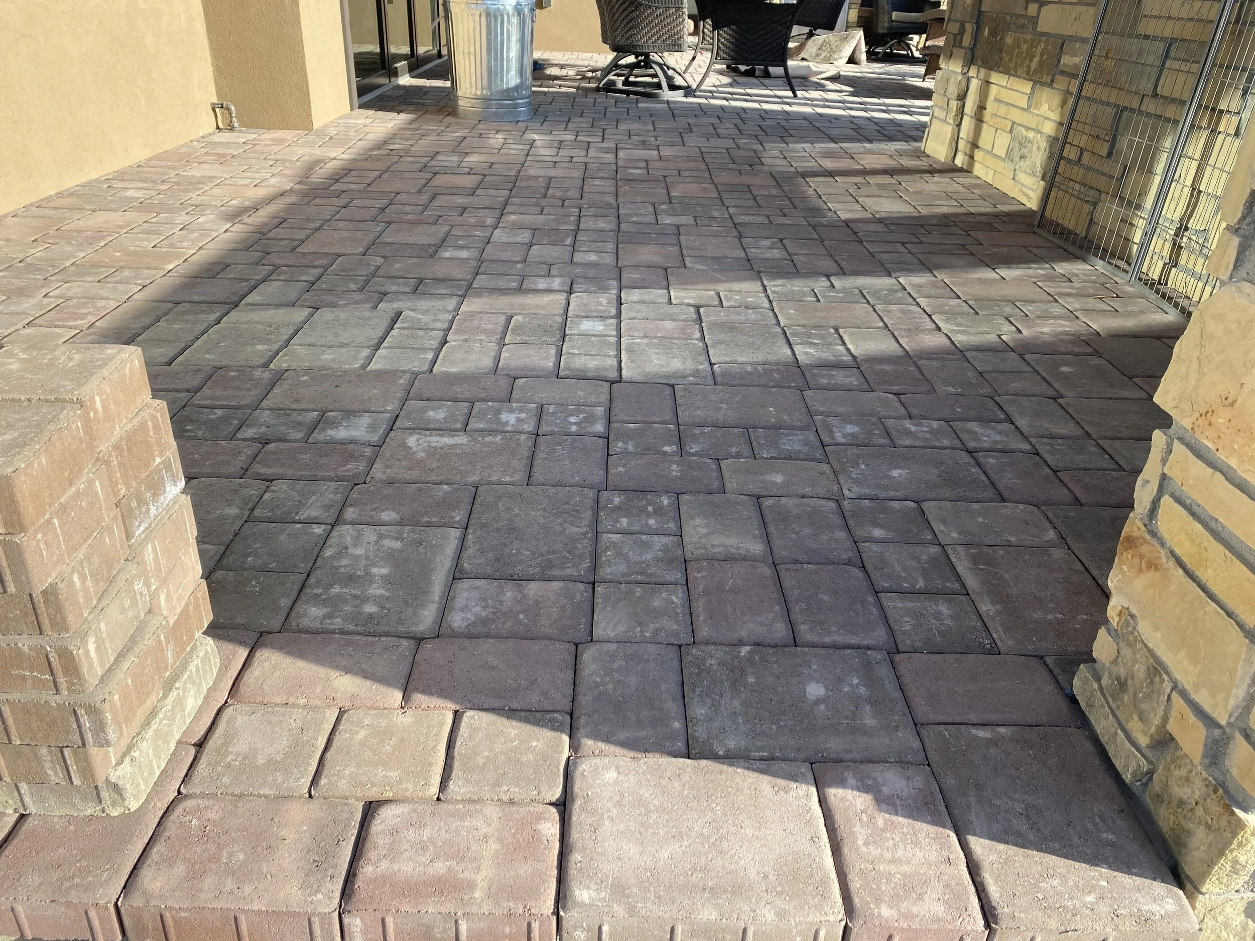 A patio paved with interlocking stone bricks or pavers in various earth tones, leading to an outdoor seating area with chairs and a table in the background, adjacent to a building with brick and stone walls.