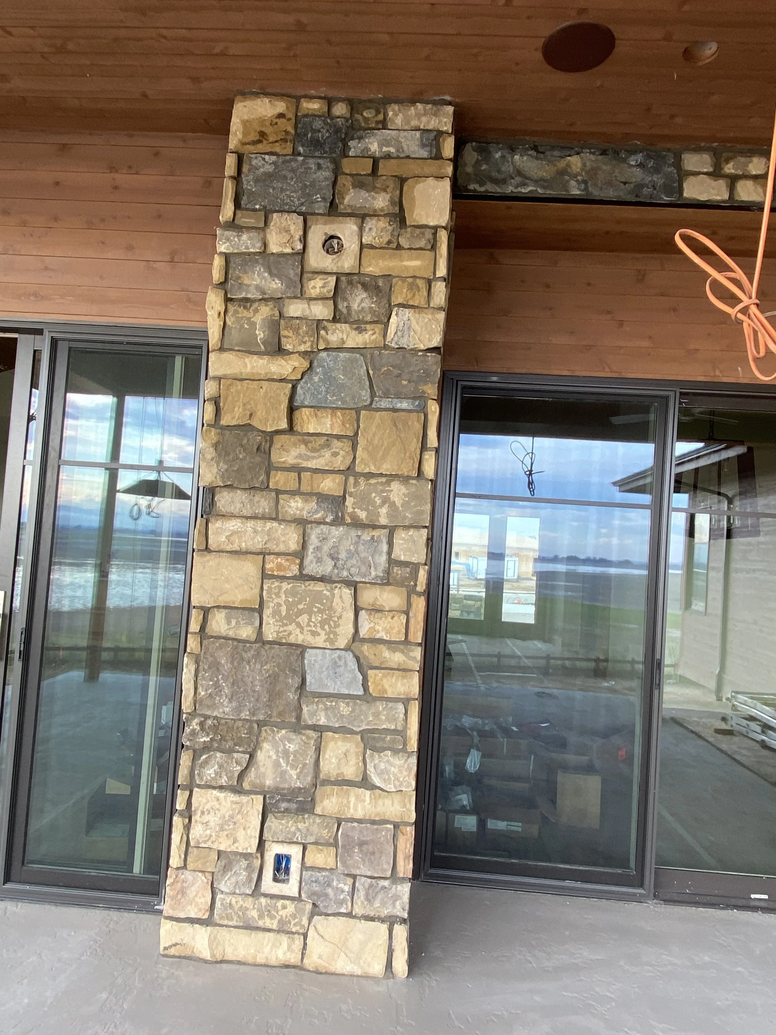 Exterior view of a building under construction, featuring a stone pillar between large glass sliding doors, with a wooden ceiling and unfinished electrical wiring visible.