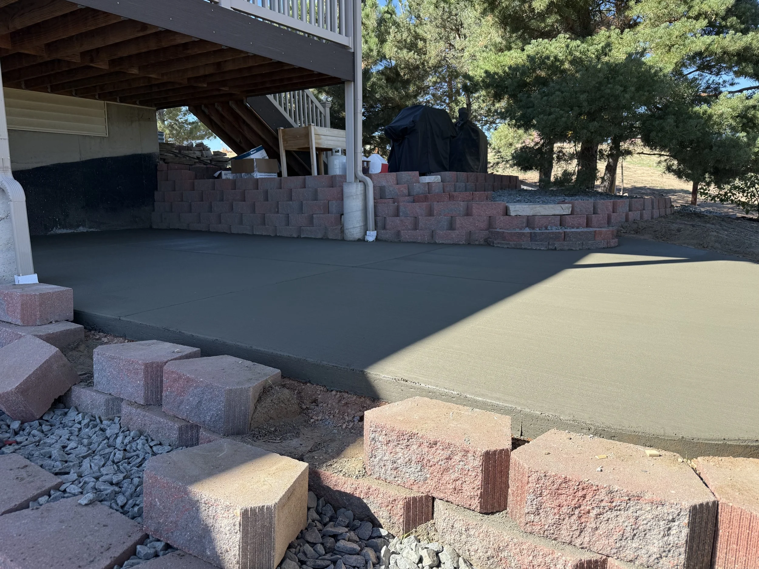 Freshly poured concrete slab partially covered by a house with a sliding board staircase leading to the upper level. Red bricks forming a retaining wall and a staircase surround the slab. Trees and clear sky are visible in the background.