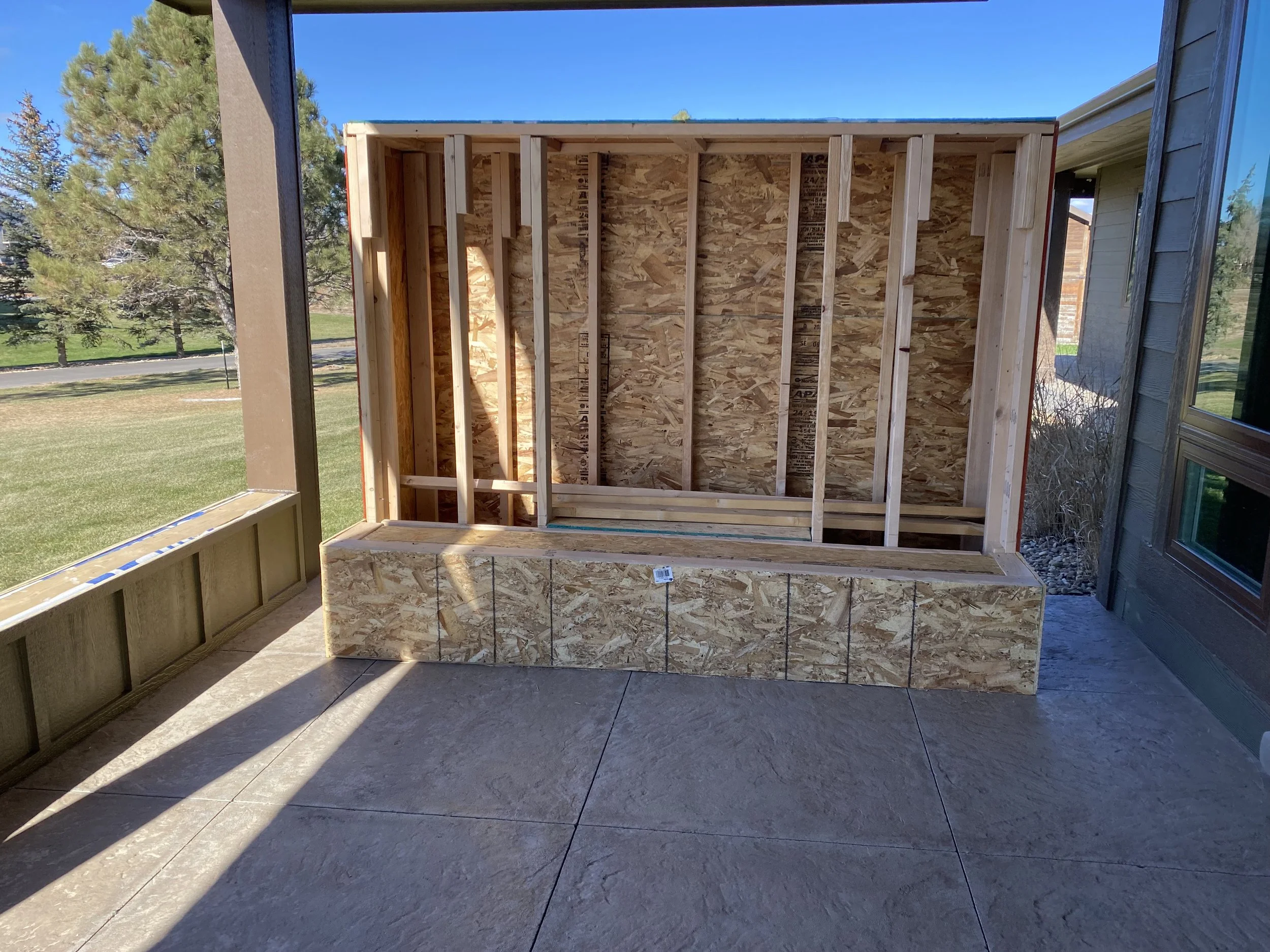 Frame of a small wooden structure under construction on a patio with a grassy backyard and trees in the background.