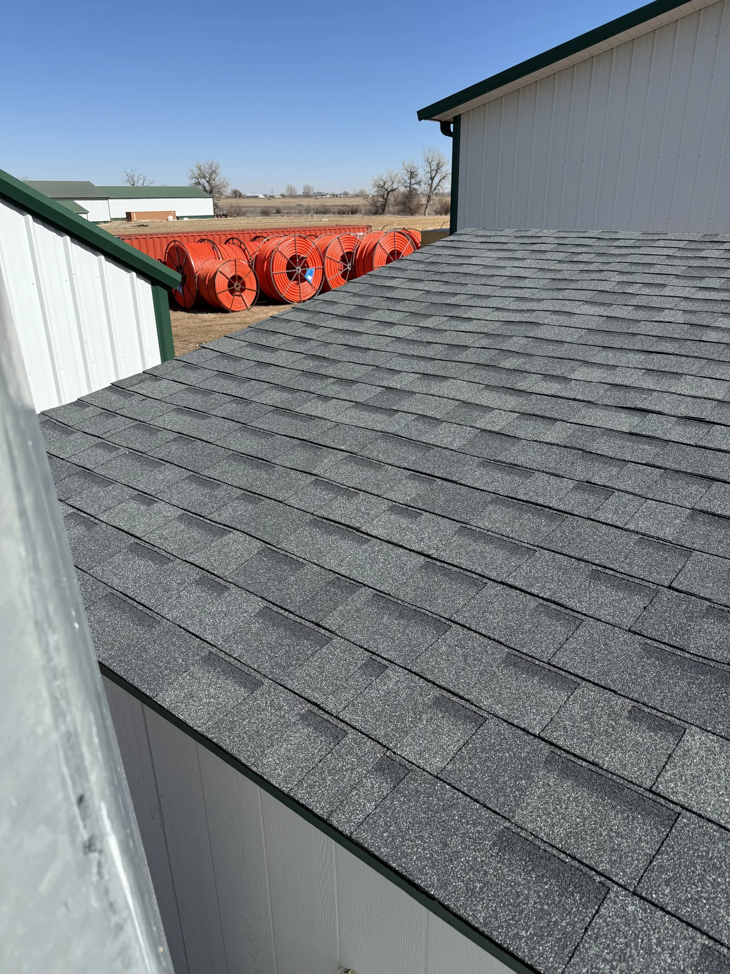 View of a gray shingle roof, with storage buildings with white siding and green trim, and orange hose spool reels in the background.
