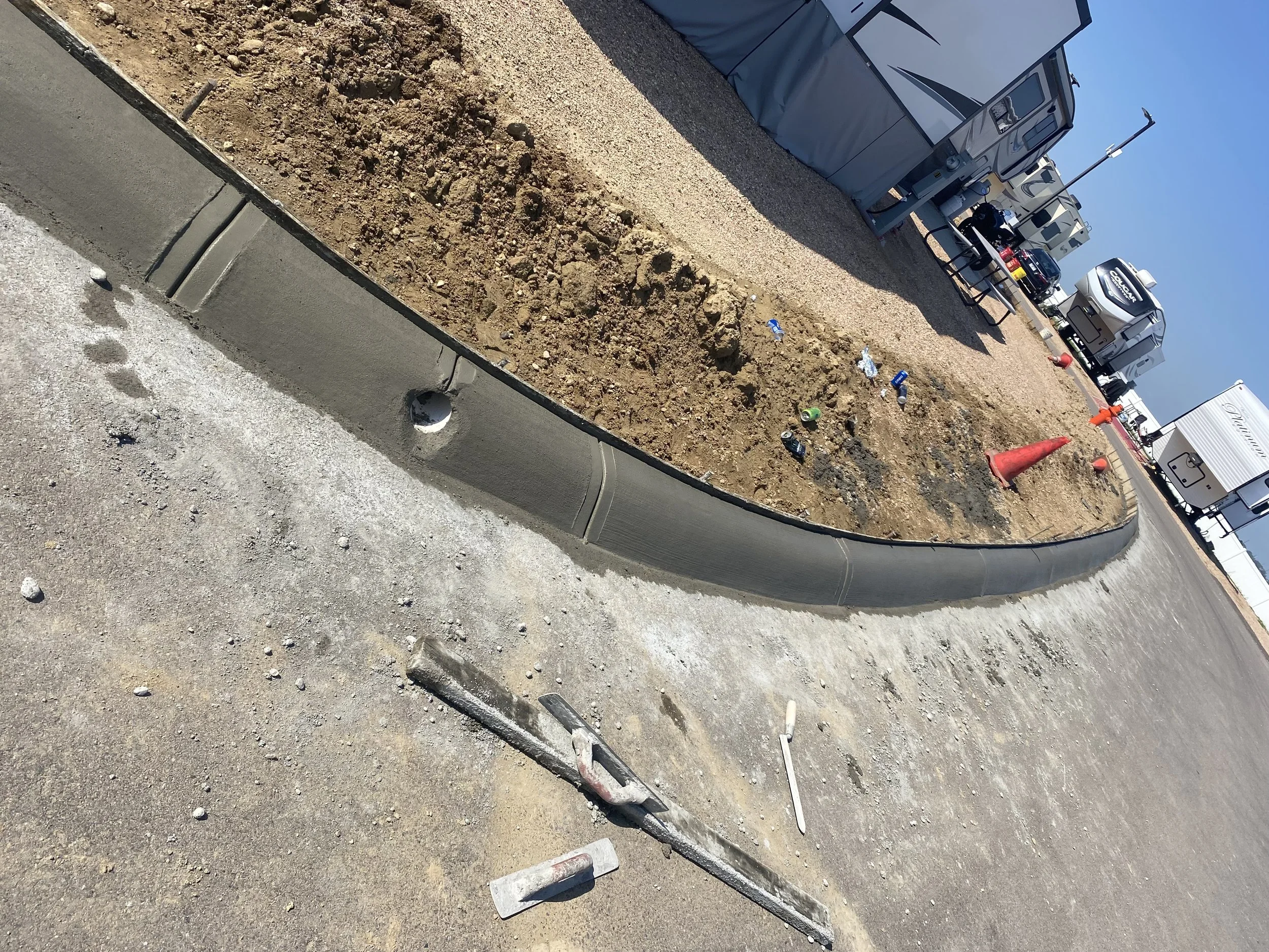 Construction scene showing a freshly built concrete curb separating a dirt area with scattered trash from a paved road. Construction tools and traffic cones are present, with RVs and trailers parked in the background under clear blue skies.