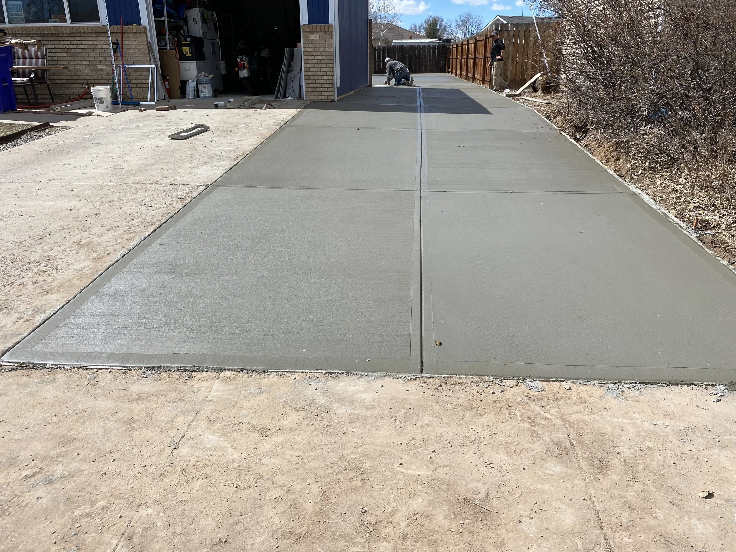 Newly poured concrete driveway in front of a garage, with two workers working on leveling and finishing the cement, and construction tools nearby.