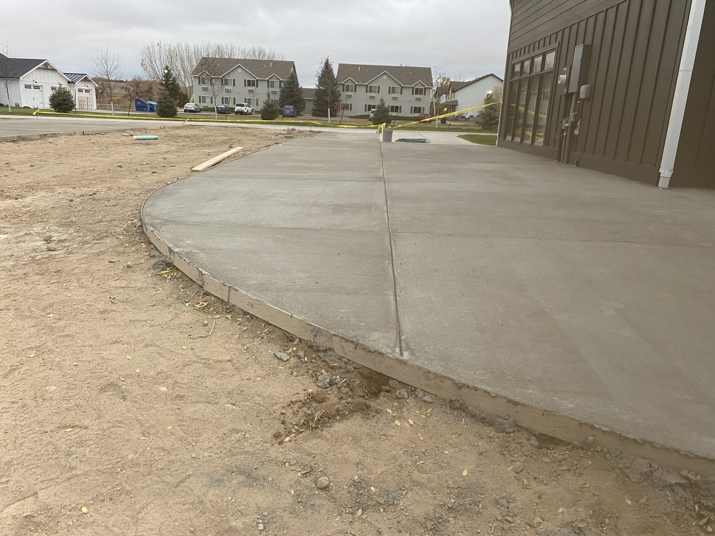 Concrete sidewalk newly poured next to building with black exterior, with dirt and construction materials nearby, in a residential neighborhood.