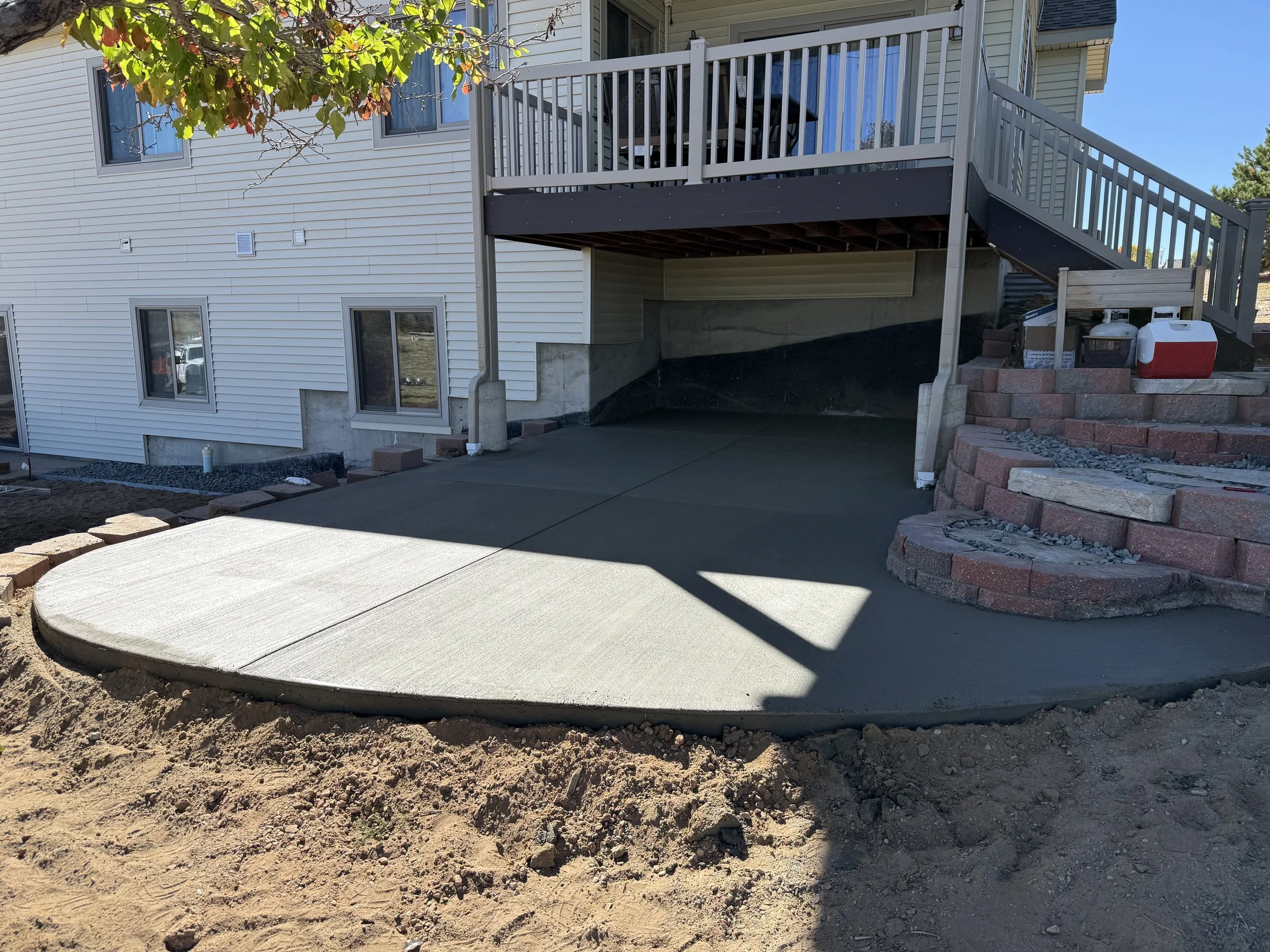 Newly poured concrete patio in the backyard of a house, with a staircase leading up to a deck and a landscaped area with bricks and rocks.