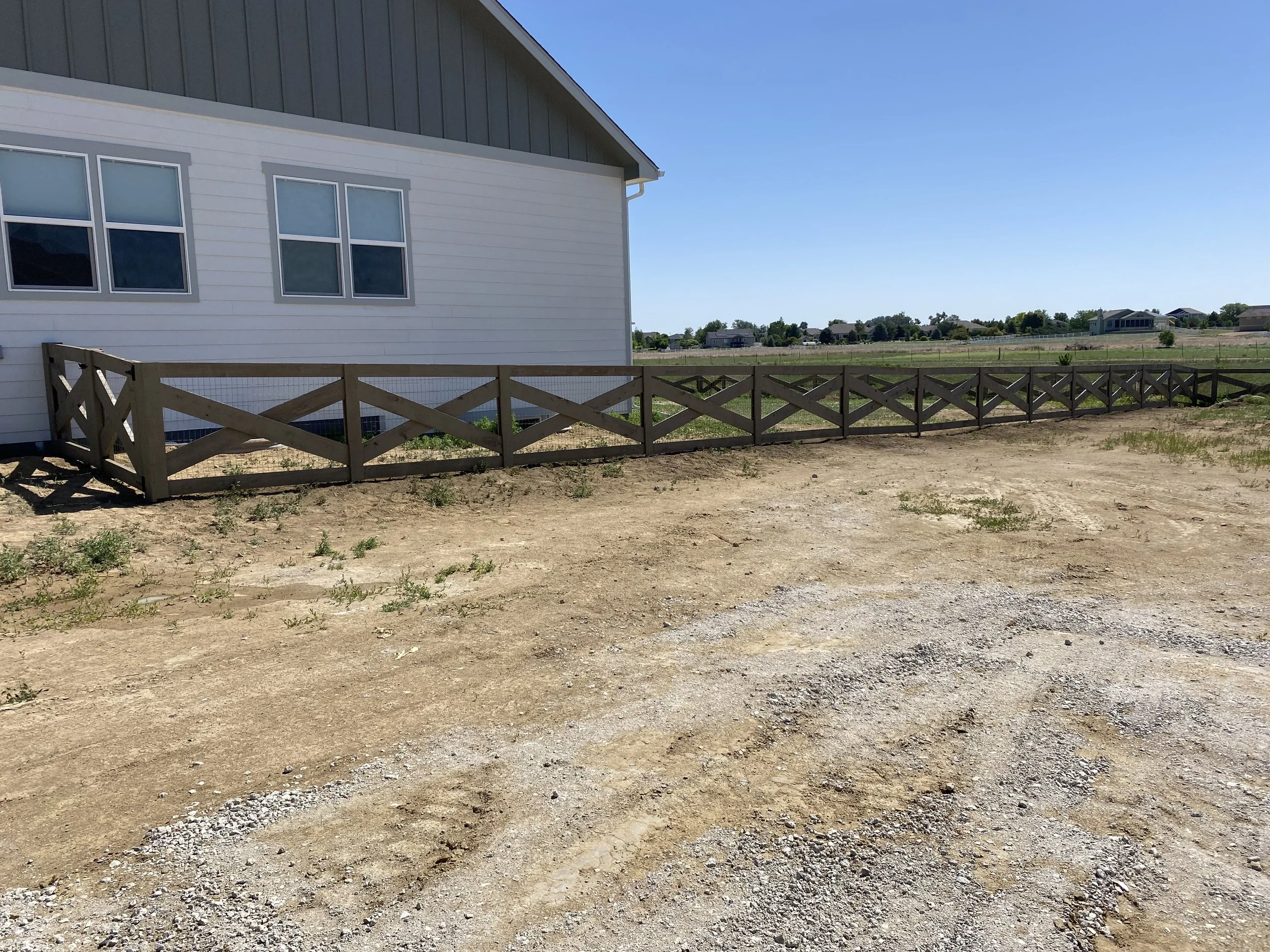 A white house with four windows, a brown wooden fence, and an unpaved, slightly bumpy yard with some sparse grass, against a backdrop of a vast open sky and distant houses.