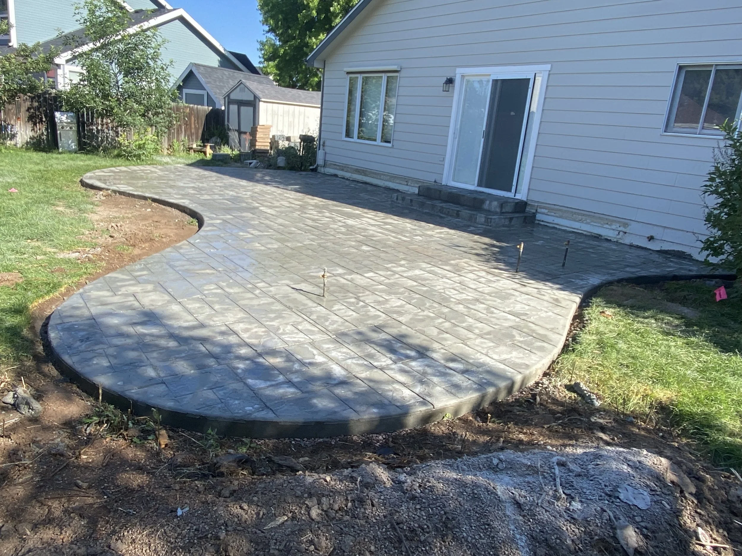 Newly paved concrete pathway in a backyard with a shrub to the right and a wooden fence in the background, leading to a house with white siding and a sliding glass door.