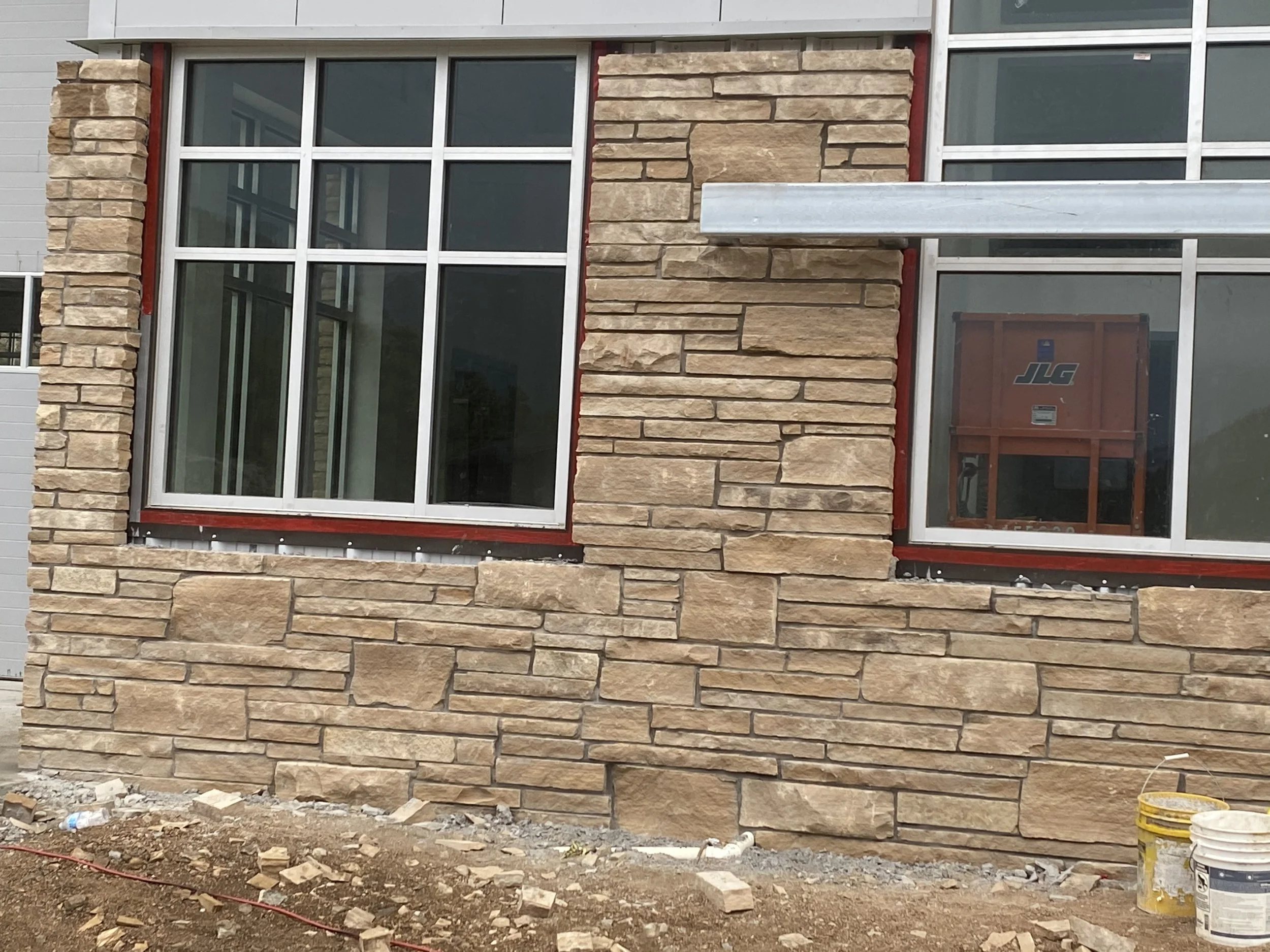 Construction site with a partially finished stone building facade and installation of large windows. Construction materials and debris are present on the ground.