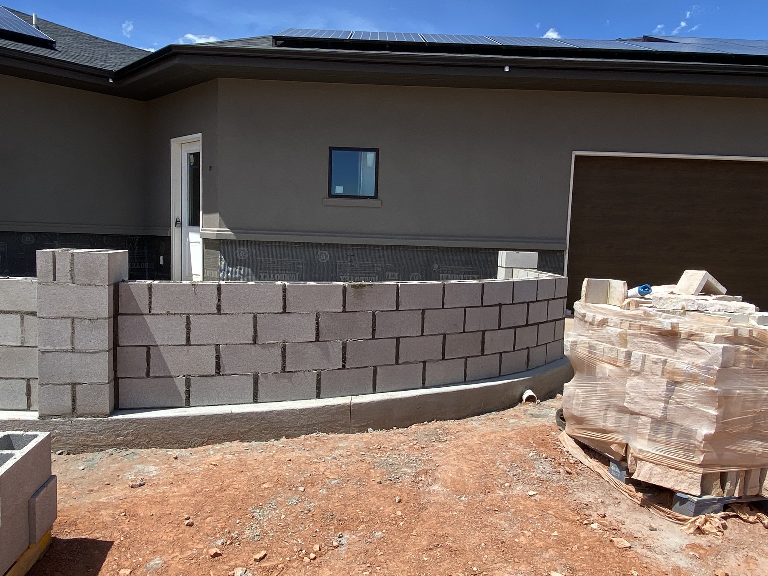Construction site of a house with a partially built curved brick wall in front of a beige house with a small window and a door, and a brown garage door to the right. Piles of bricks wrapped in plastic and construction materials are on the ground.