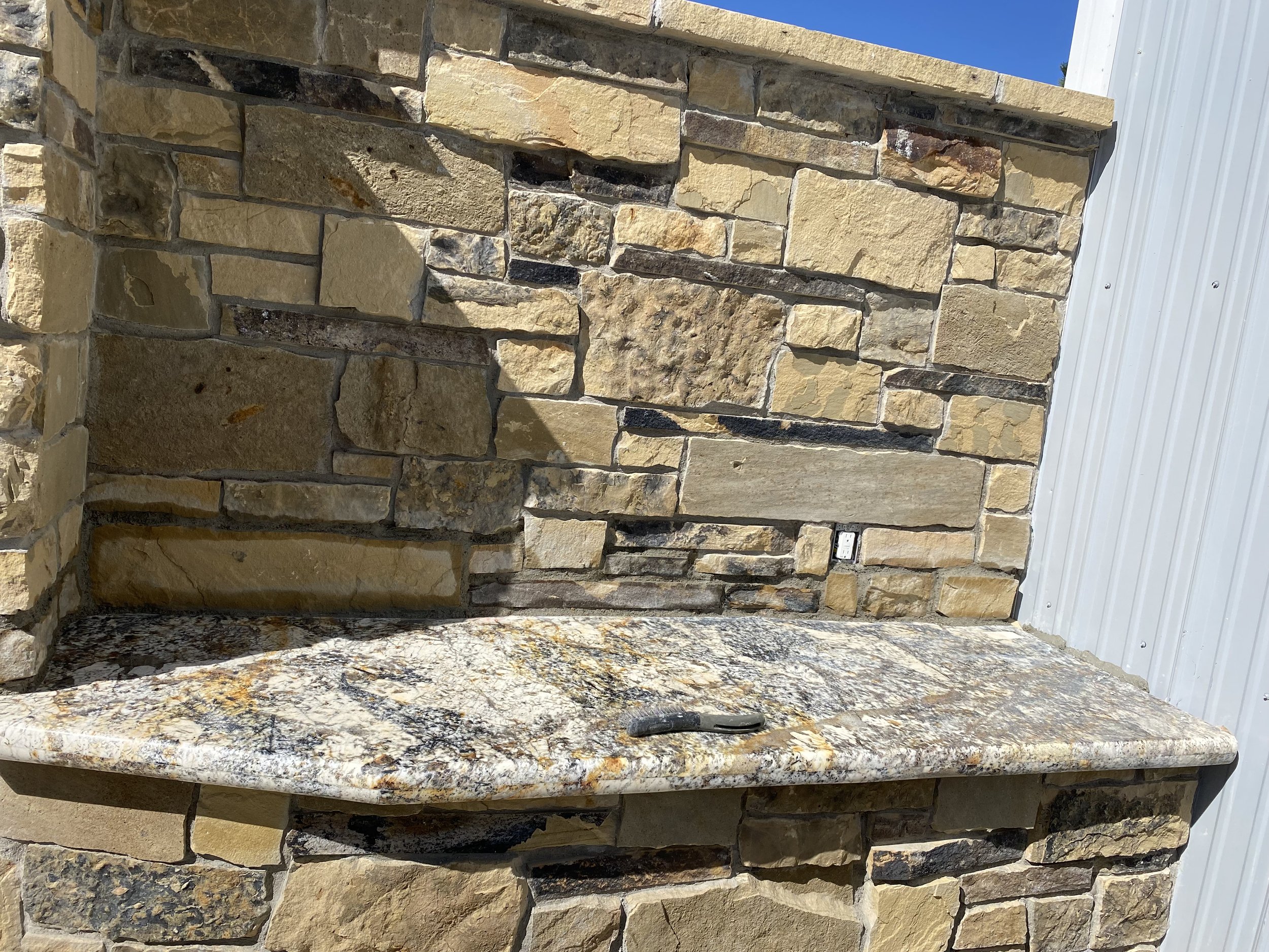 Close-up of a stone outdoor countertop with a multi-colored granite surface, set against a stone wall with a small black tool placed on it.