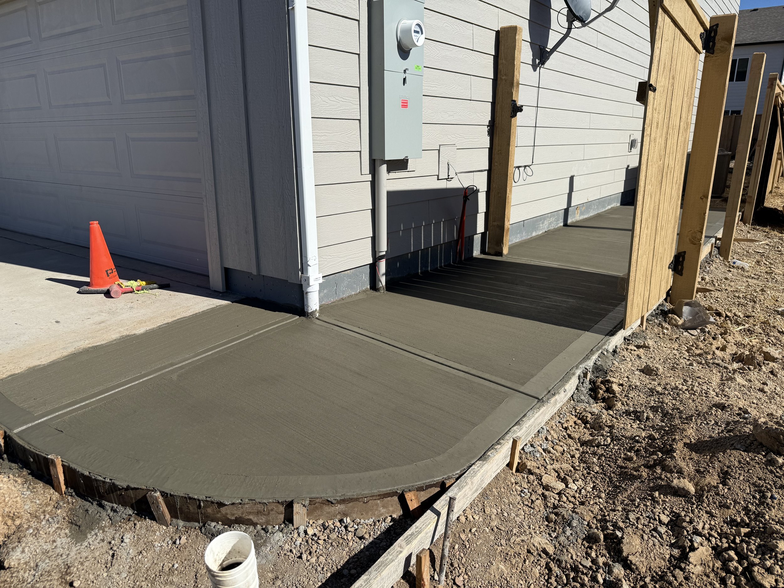 Newly poured concrete sidewalk leading to a house with a ramp, surrounded by wooden framing and construction materials.