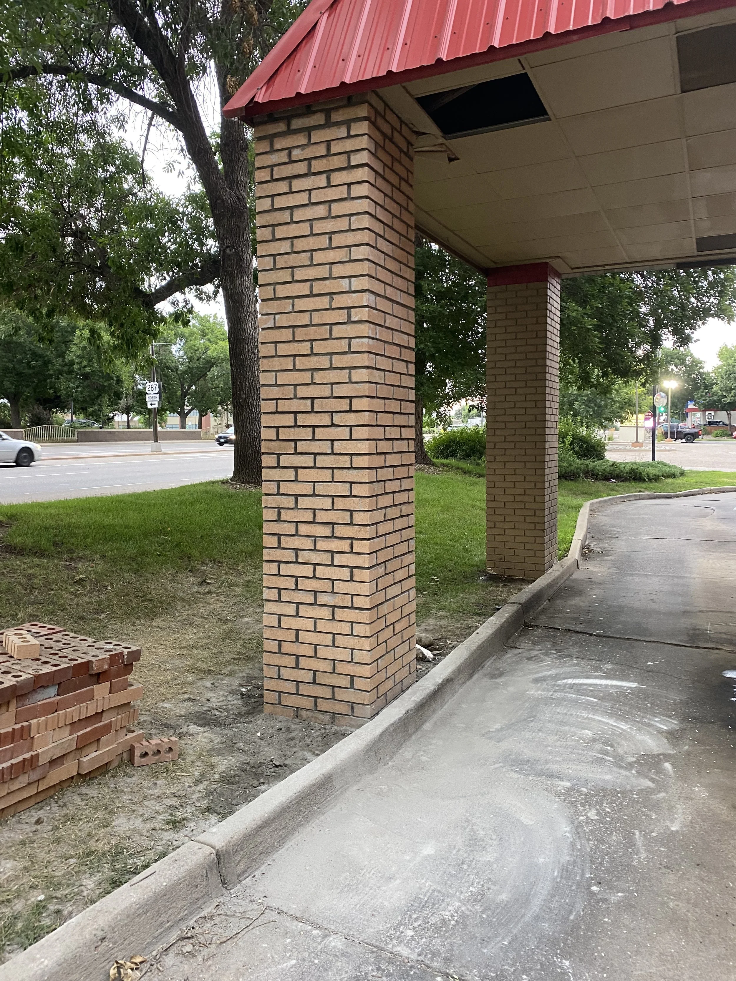 The image shows the exterior corner of a building with brick columns supporting a red metal roof. There is a sidewalk with white chalk markings on it and a small pile of unused bricks on the ground. In the background, there are trees, a road with moving cars, and some commercial buildings.