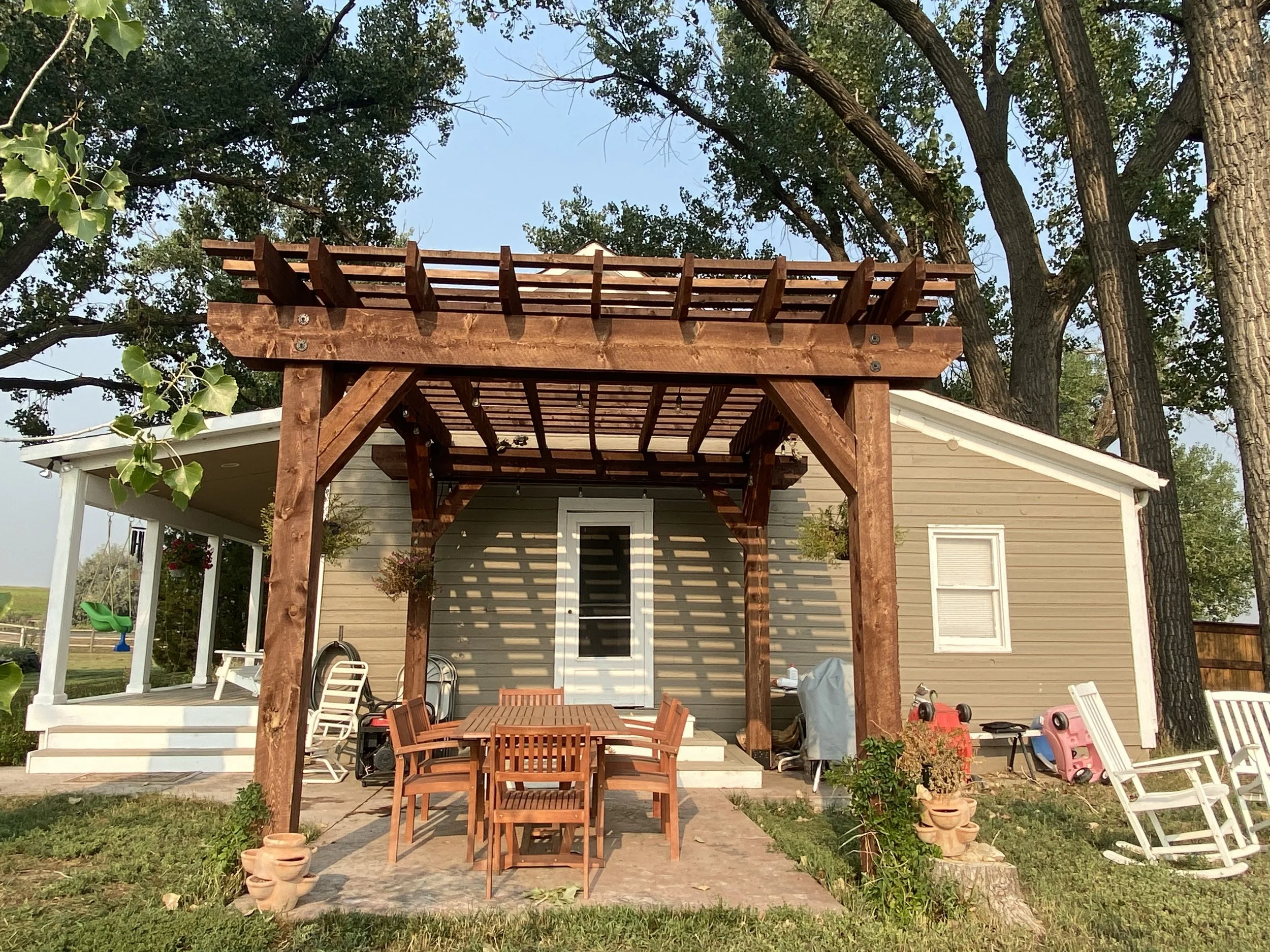 Backyard patio with wooden pergola, outdoor dining table, chairs, and swing set in a suburban backyard.