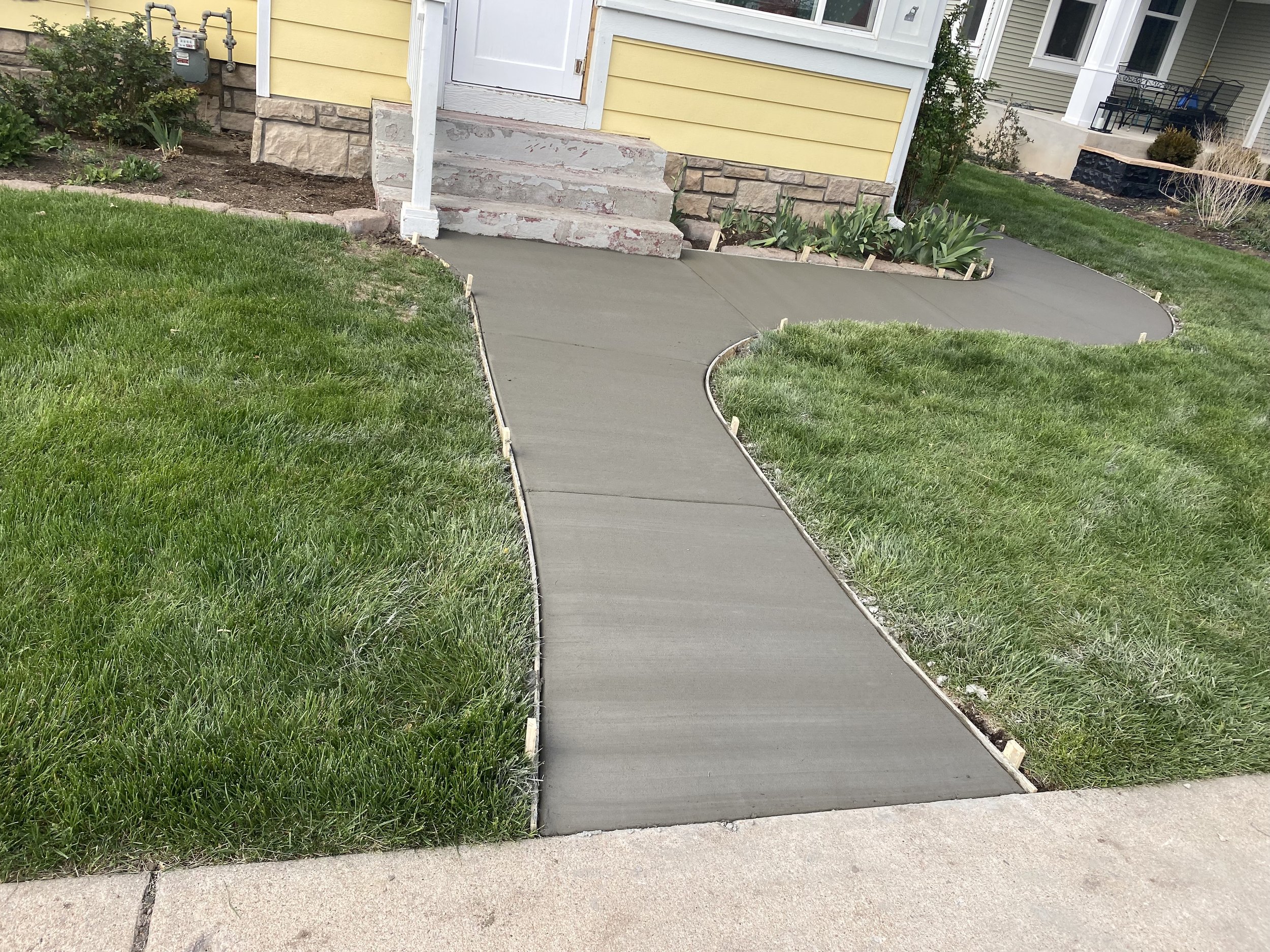 Newly poured concrete sidewalk curving towards the front entrance of a yellow house with stone foundation, surrounded by green grass and flower beds.