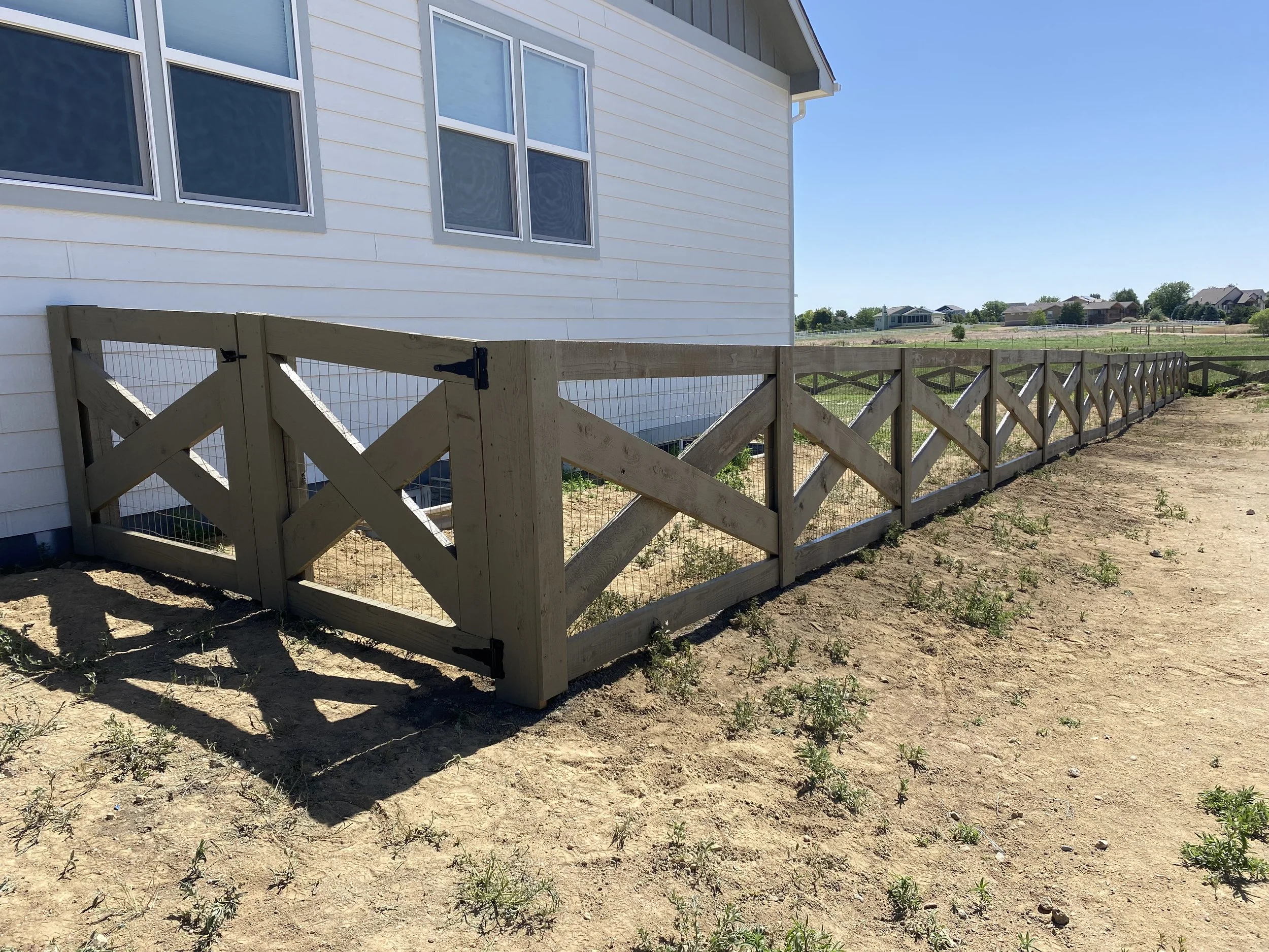 A wooden fence with wire mesh runs alongside a white house with multiple windows on a sunny day.