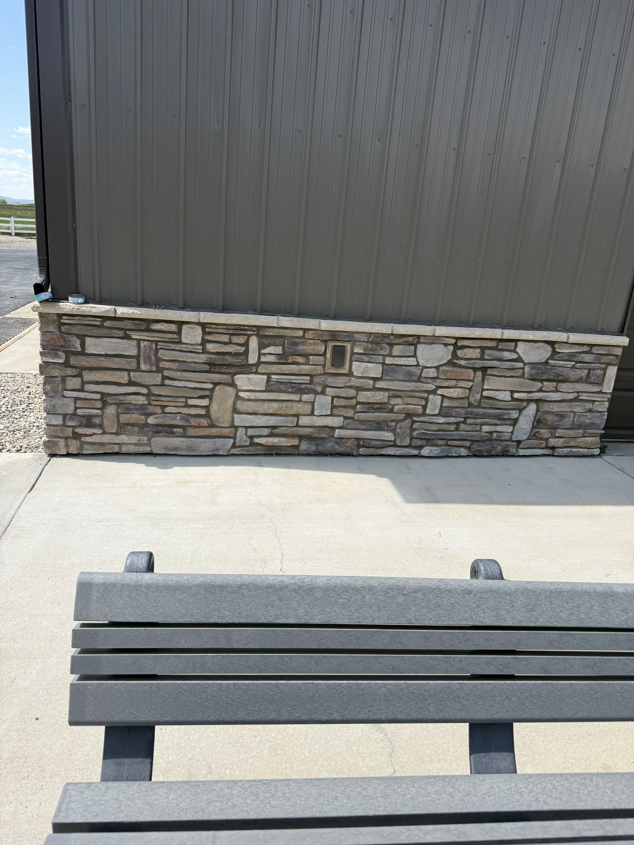 Empty park bench on concrete pavement in front of a building with a stone and metal wall and clear sky in the background.