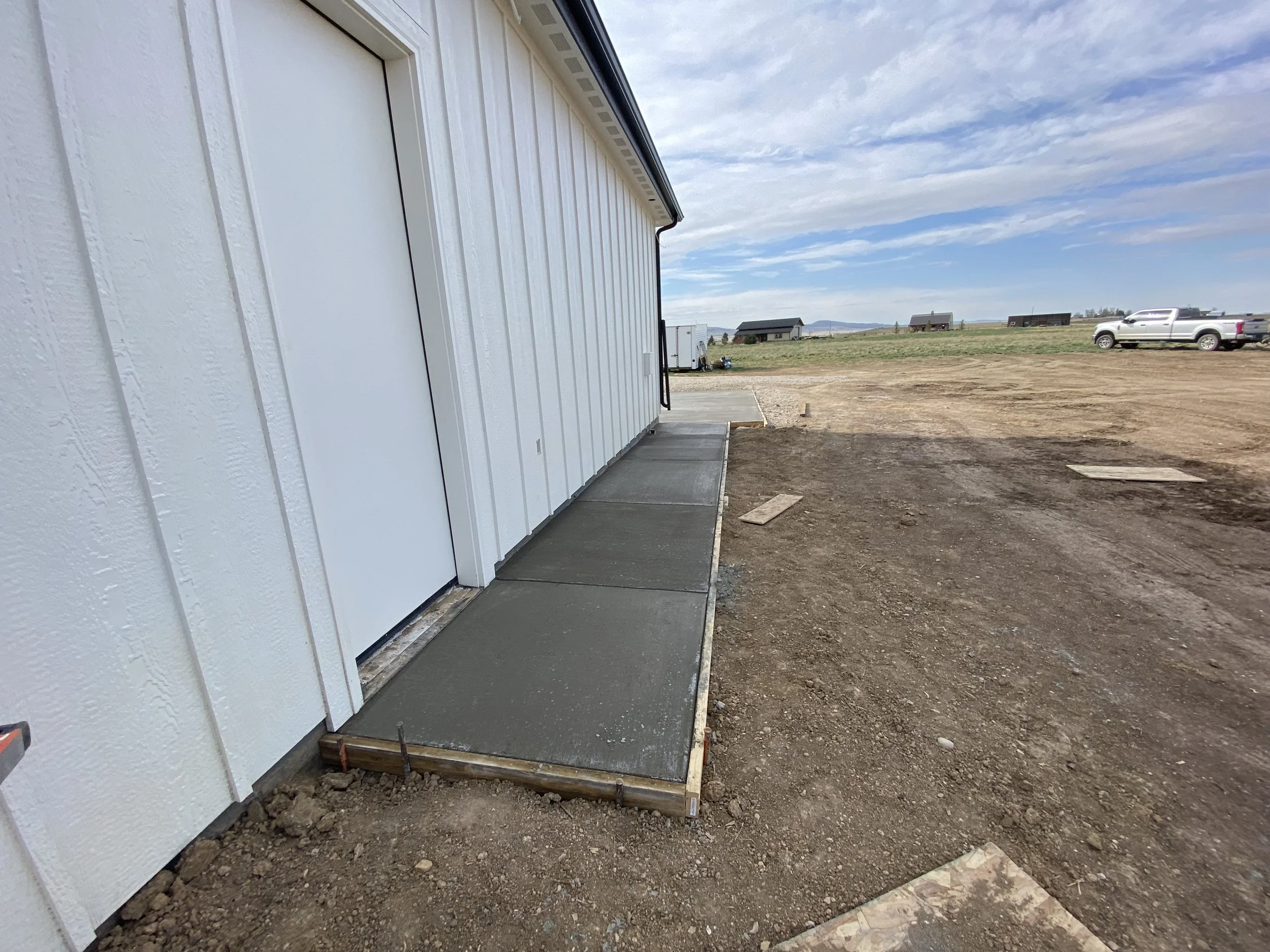 Newly poured concrete sidewalk and foundation at the side of a white building in a rural area with trucks and small buildings in the background on a partly cloudy day.