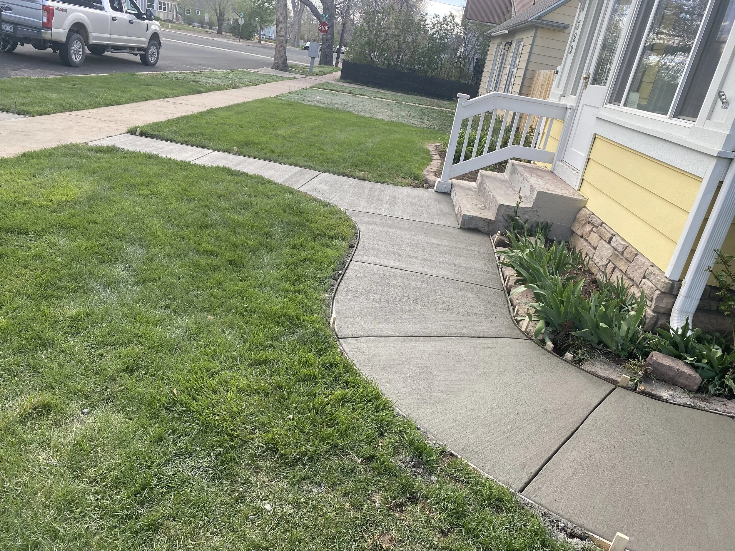 Concrete sidewalk curving in front of a yellow house with white trim, small steps leading to a porch, and a garden bed with green plants, next to a well-maintained lawn.