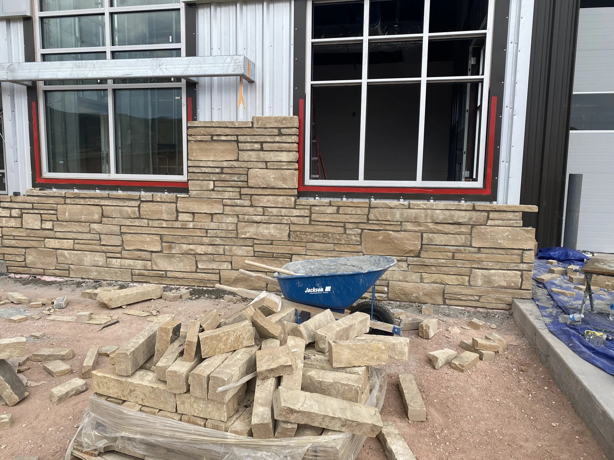 Construction site with a partially built stone wall in front of a building with window and black siding. Piles of stone bricks and a blue wheelbarrow are on the ground.