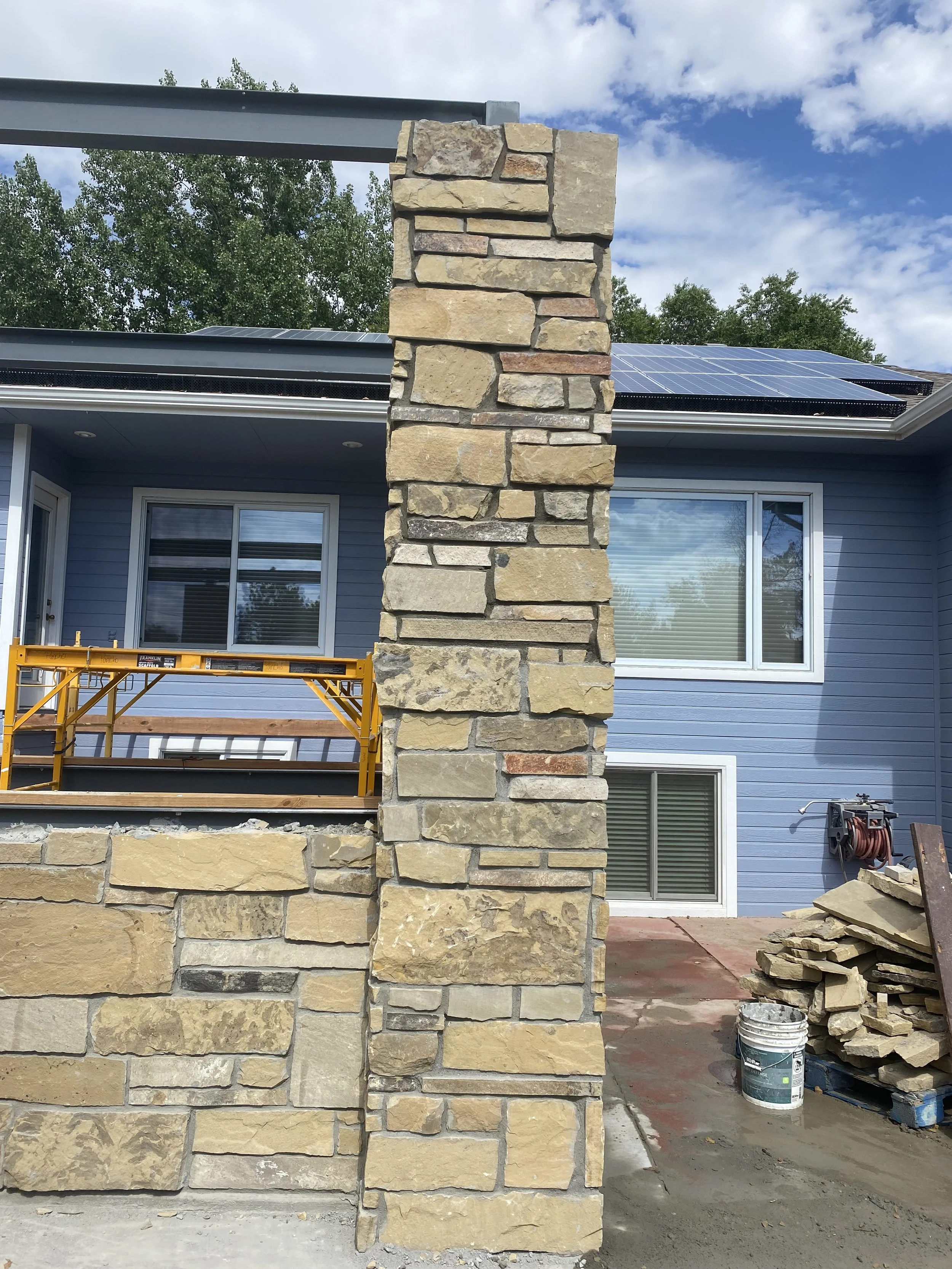 A house under construction with a stone pillar wall in front. The house has blue siding, large windows, and solar panels on the roof. Construction materials and tools are visible beside the house.