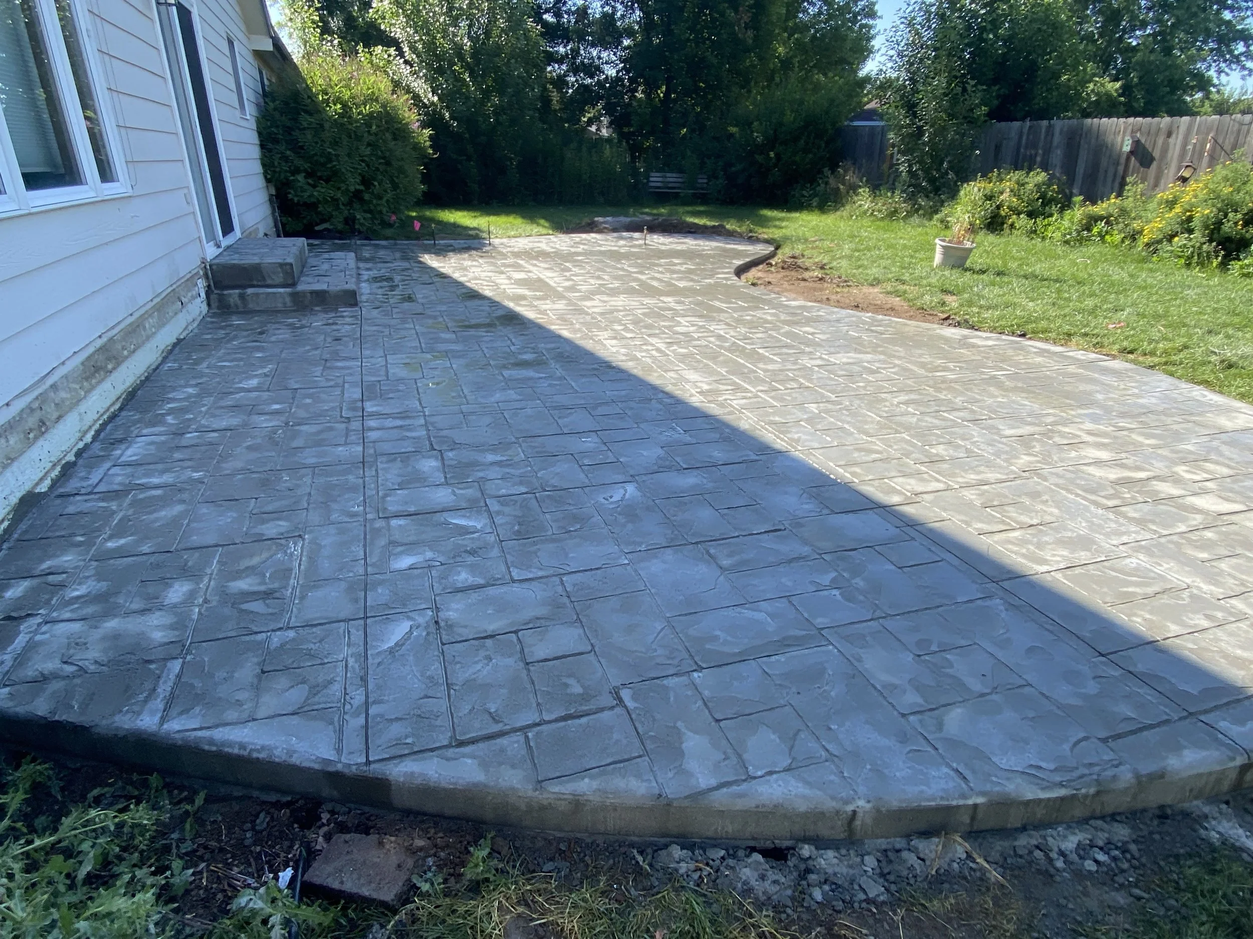 Newly paved concrete patio with stamped stone pattern next to a house with white siding, surrounded by green shrubs and a fenced yard.
