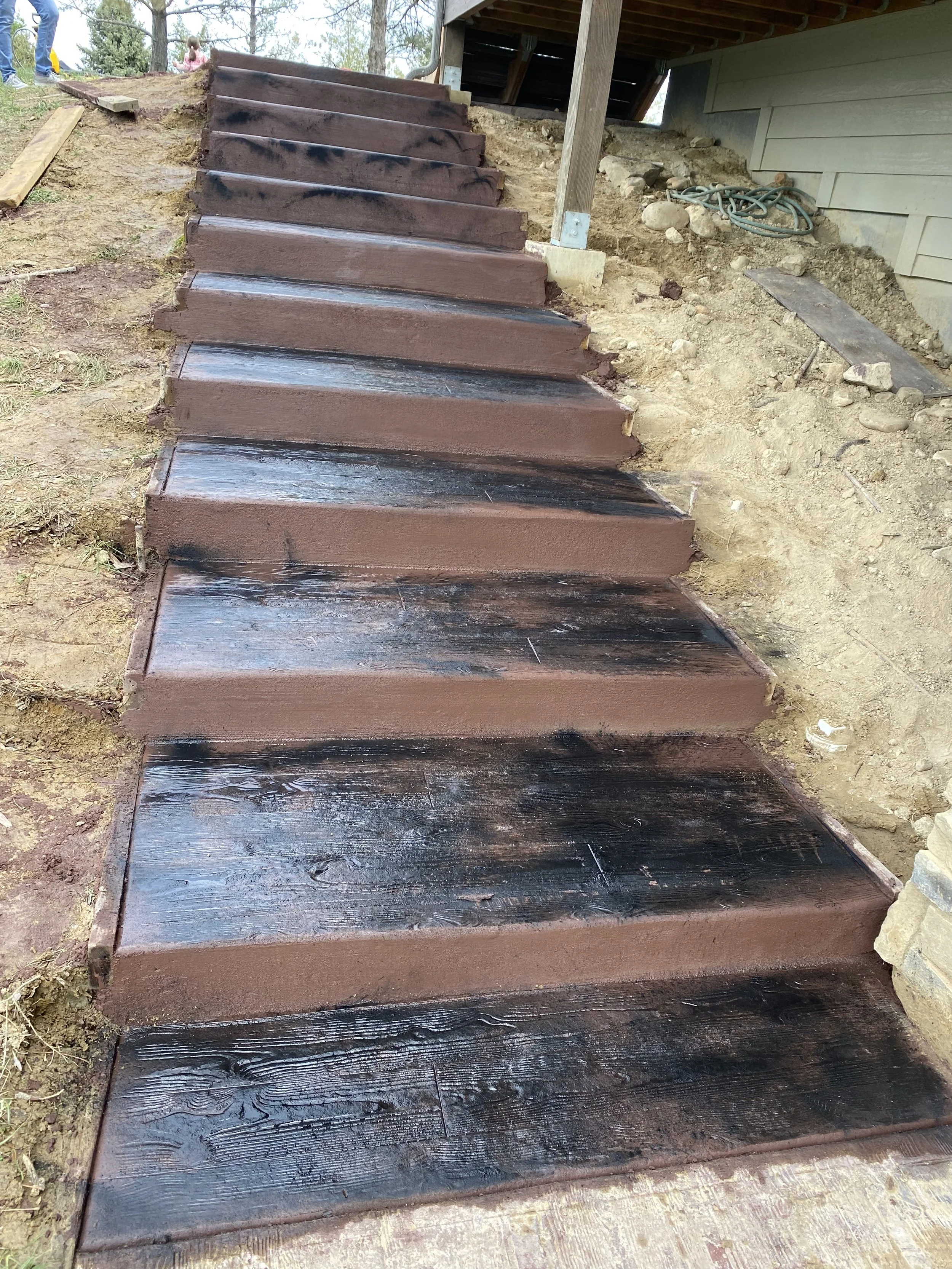 Newly constructed outdoor wooden stairs under a porch, with some sections freshly painted dark brown and others still showing wood grain, leading up to a yard with children playing.