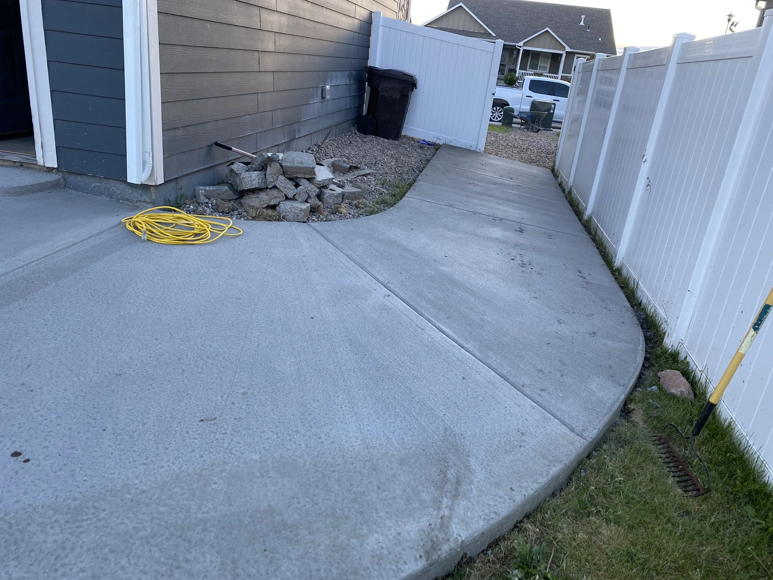 A concrete driveway with a curved section leading to a gate, surrounded by a white fence, a gray house wall, a pile of rocks, a yellow garden hose, and a rake, with a backyard and parked vehicles visible in the background.