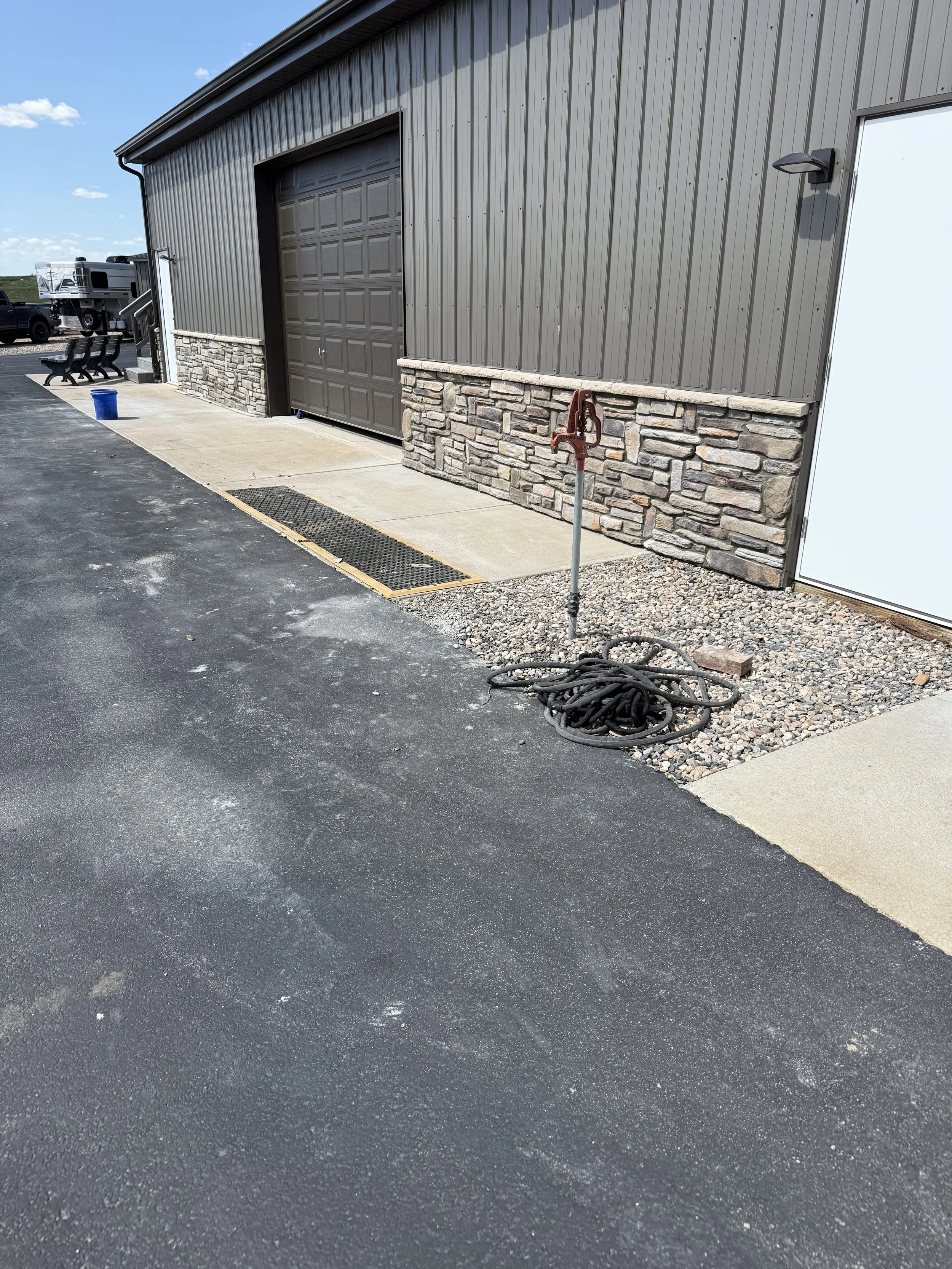 Exterior view of a building with a metal siding and stone base, a large closed garage door, and a white door. There is a fire hydrant with a hose reel nearby, a black paved surface, and some parked vehicles in the background. The scene is sunny with a clear blue sky.