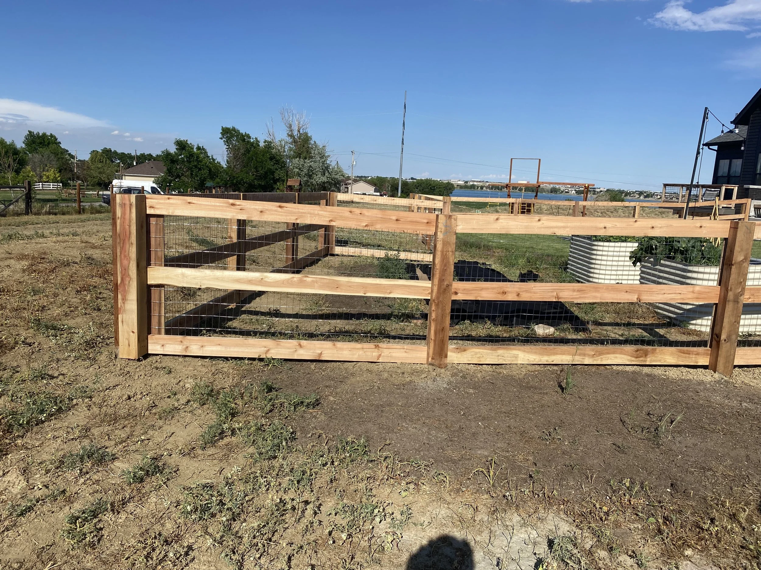 A wooden fence with wire mesh sections surrounds a dirt area with some weeds, on a sunny day with a clear blue sky, near a residential area with houses and trees in the background.