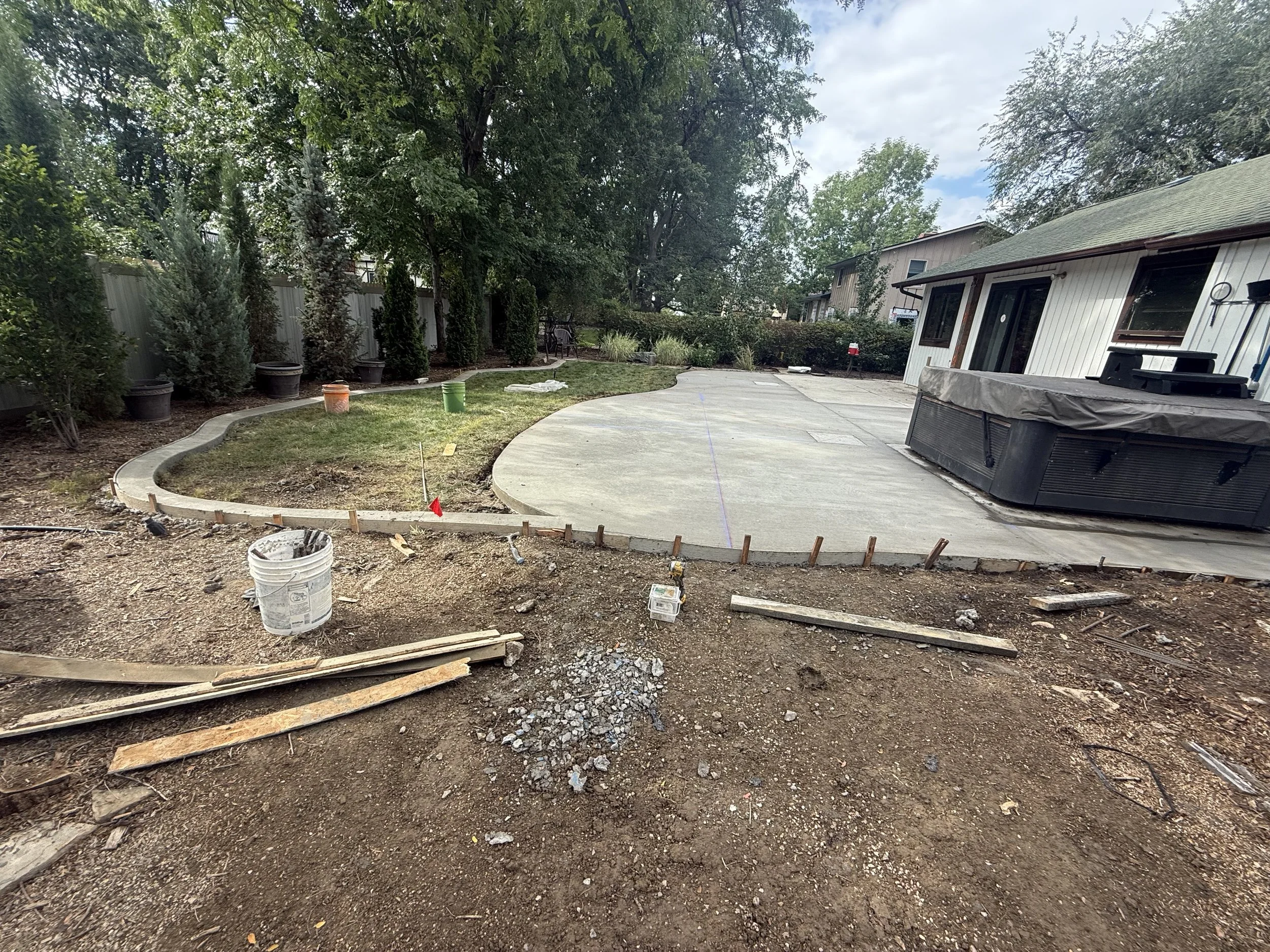 A backyard under construction with a newly poured concrete patio, bordered by a curved stone edging, and a small grass area to the left. Construction materials and tools are scattered in the foreground, and trees and a house are visible in the background.