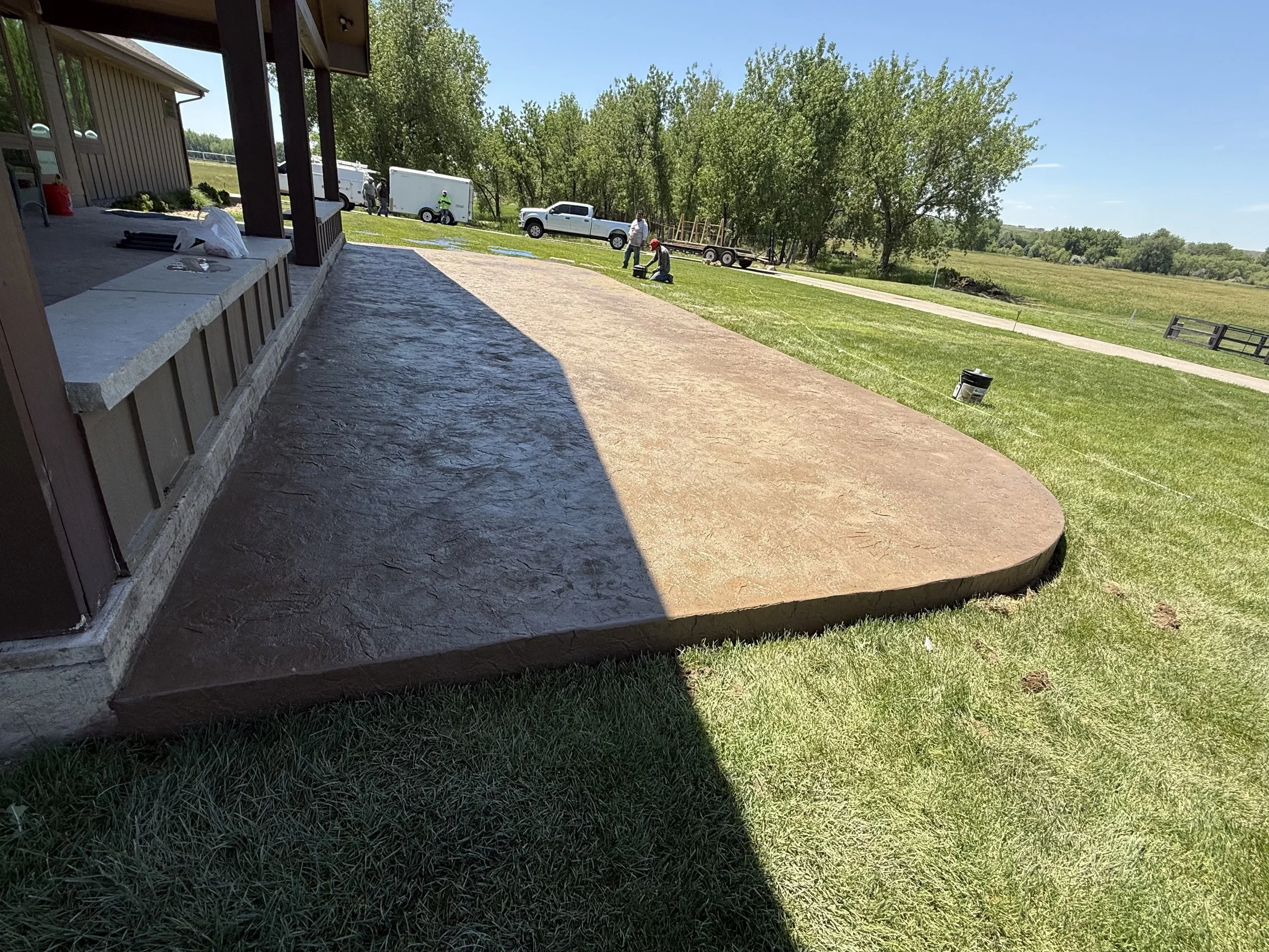A newly poured concrete patio in front of a building with a covered porch, surrounded by grassy yard and trees, with vehicles and people working in the background on a sunny day.