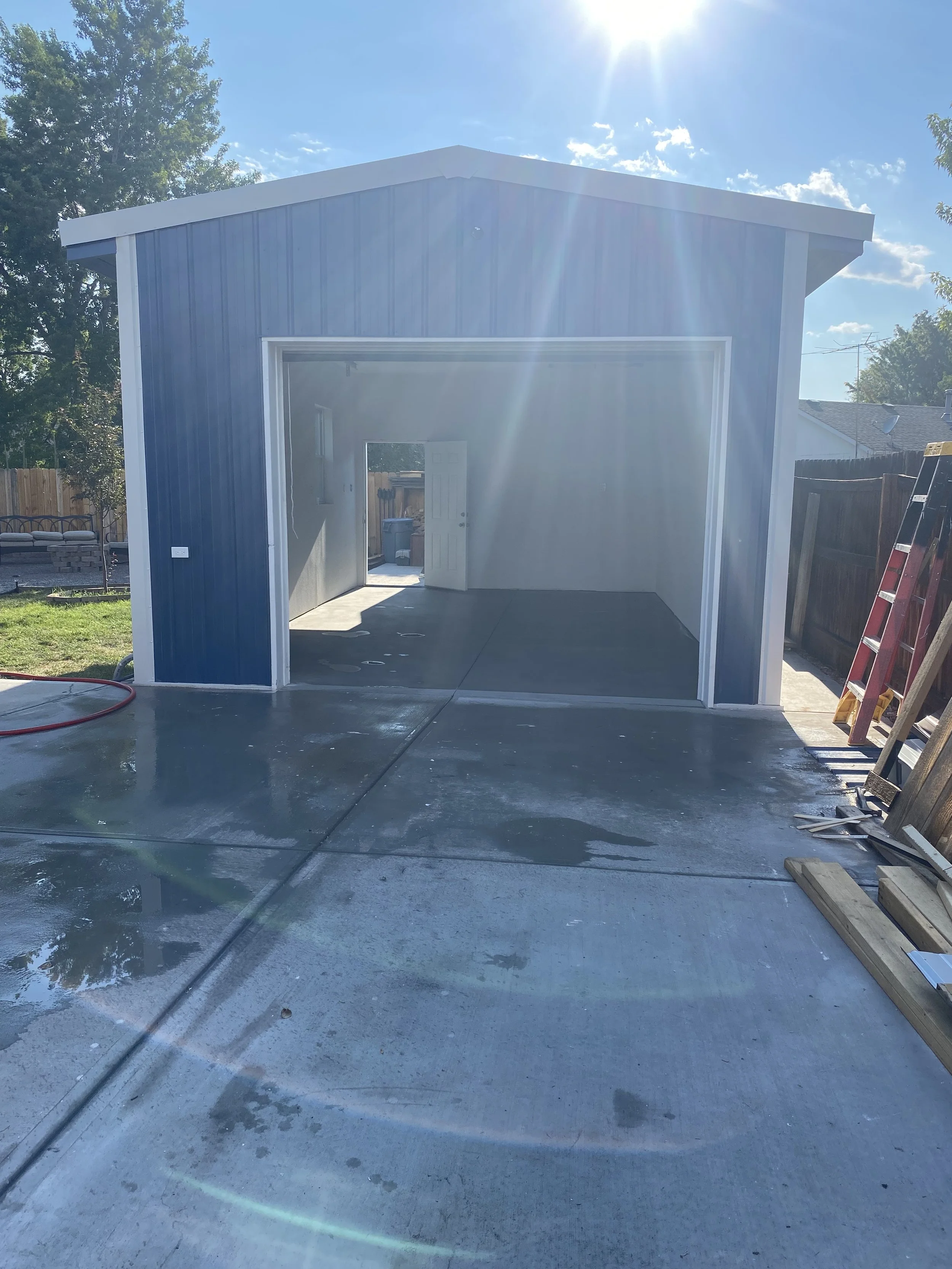 Photo of a newly built garage with blue exterior, open door, and a concrete driveway with some water patches, sunlight shining overhead, and a ladder and construction materials on the right side.