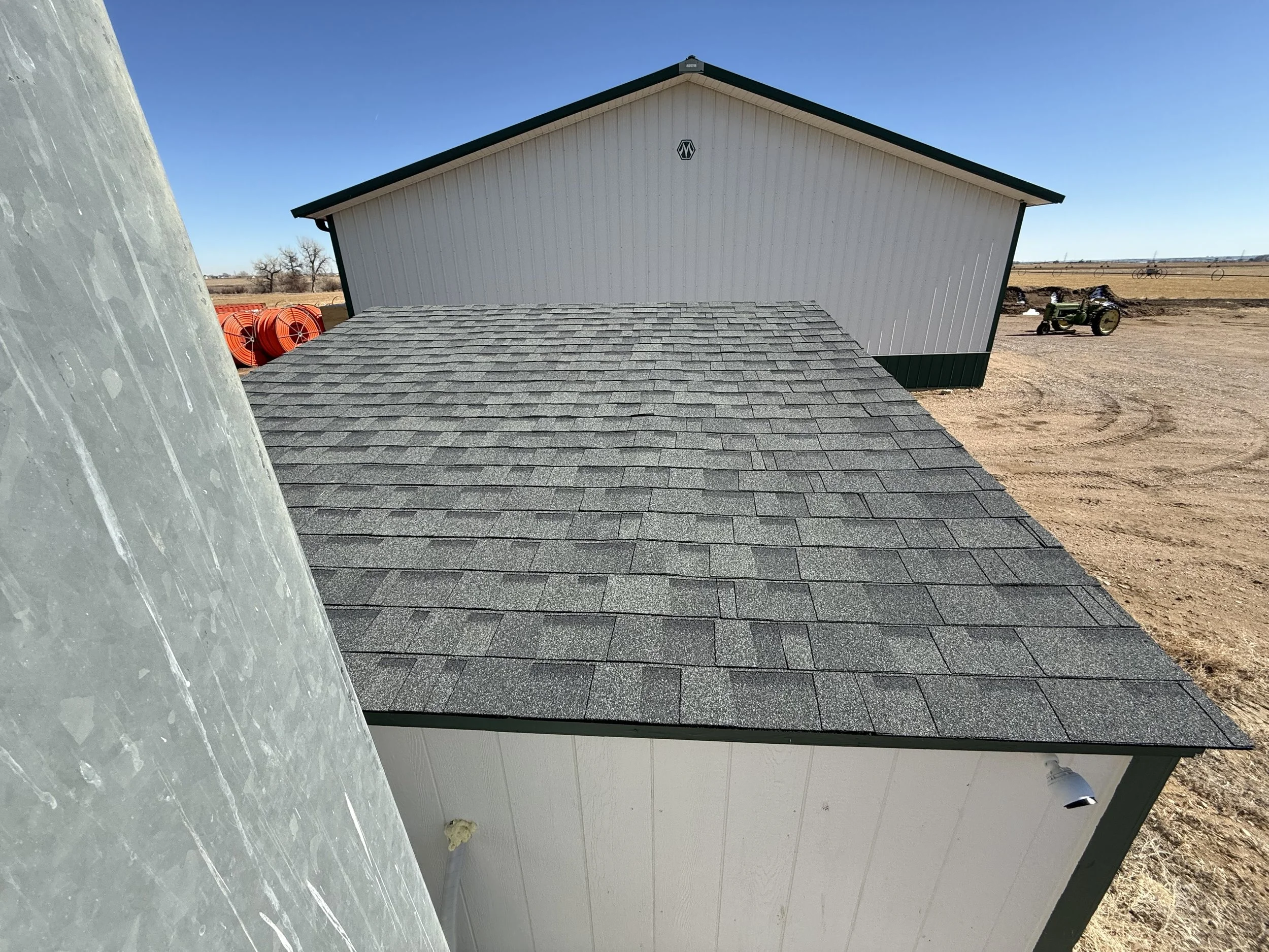View of a building with grey shingle roof, white exterior walls, and a dark green trim in a rural area with dirt ground, a tractor and orange pipes in the background, and a clear blue sky.