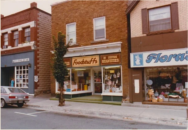 A street view showing small shops, including a store named 'foodstuff's' that sells candies and chocolates, with a small tree in front and a parked gray car on the street.