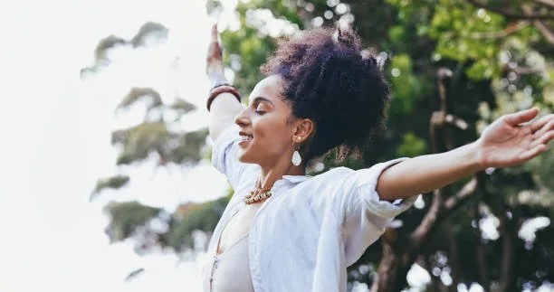 A woman with curly hair smiling and with arms outstretched outdoors among trees.