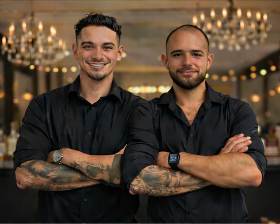 Two smiling men with arms crossed standing in a warmly lit indoor setting, possibly a restaurant or event space, dressed in black shirts with visible tattoos on their arms.