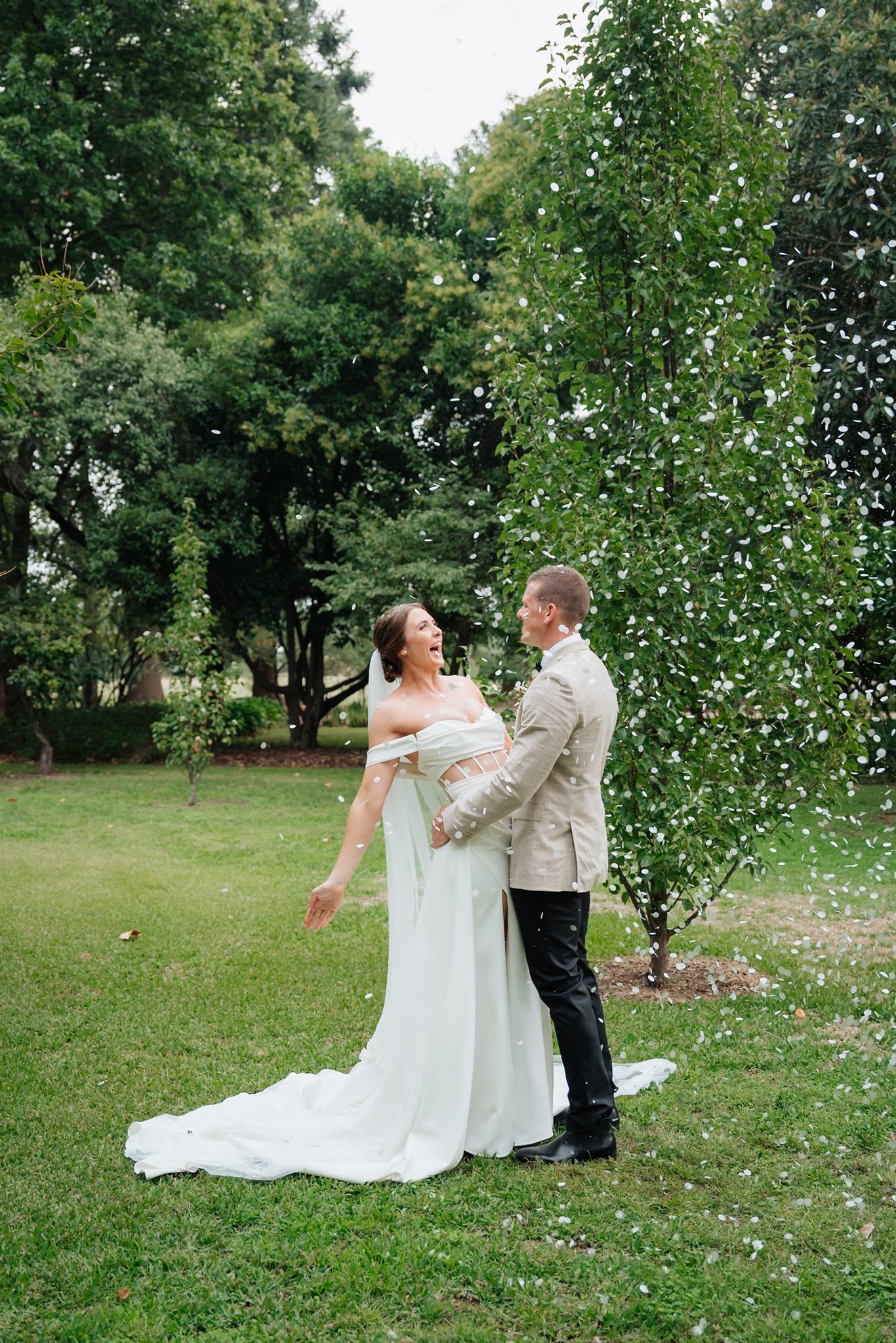 A bride and groom celebrating outdoors with white confetti falling around them, surrounded by green trees and grass.