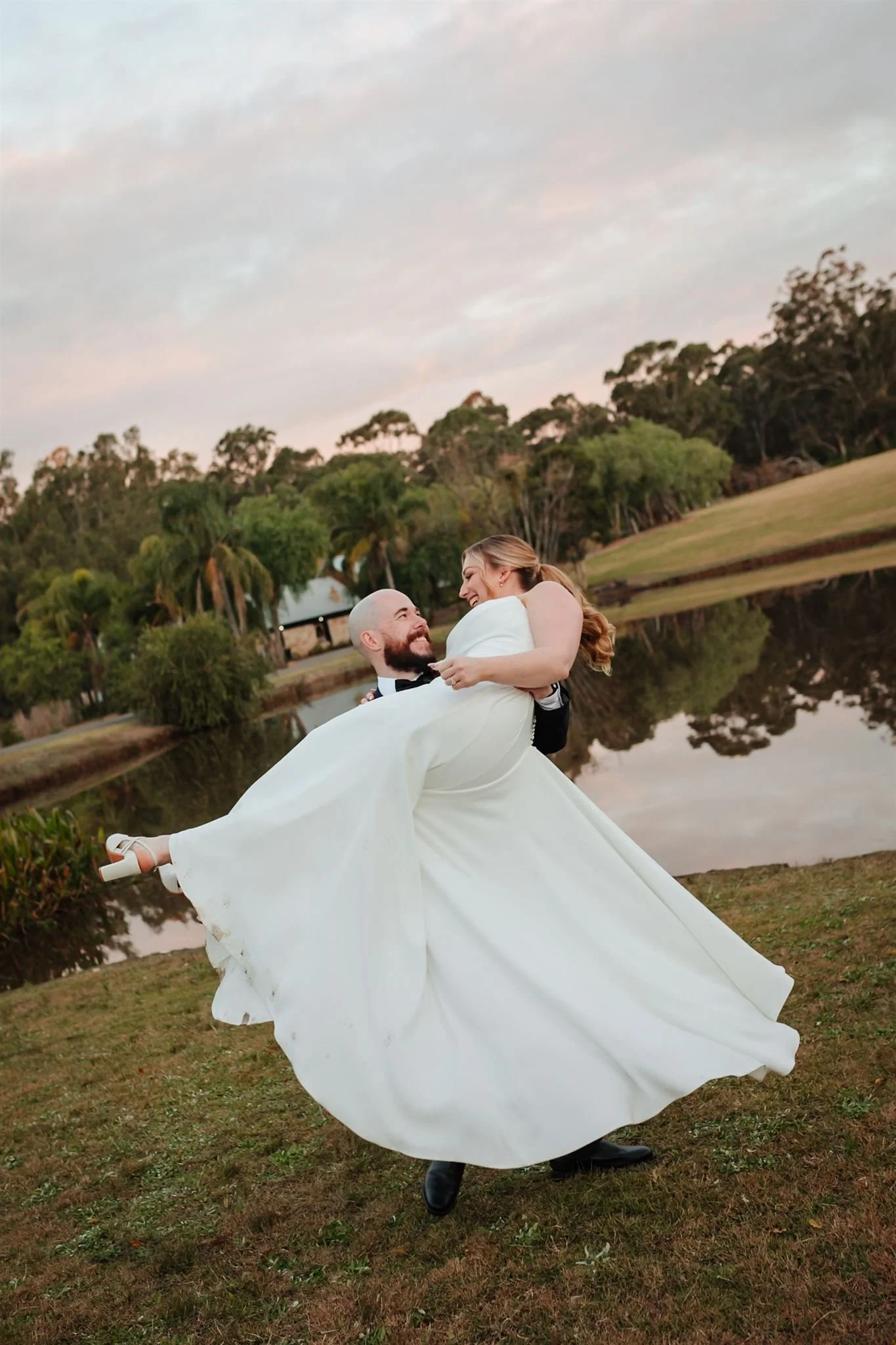 A groom and bride are dancing outdoors by a lake during sunset, with trees and a sky filled with clouds in the background.