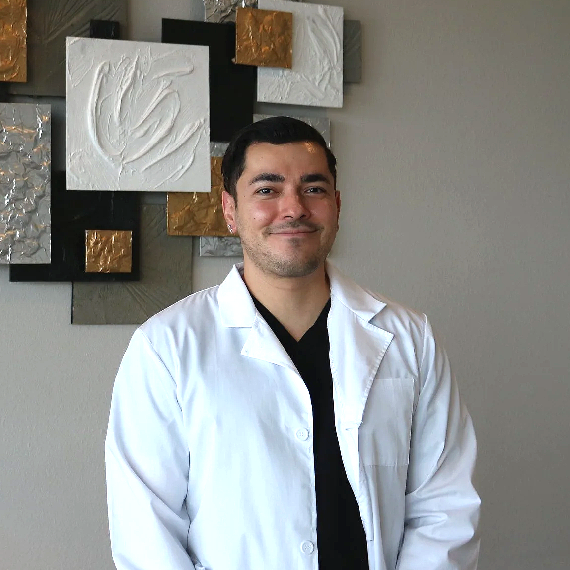 A man wearing a white lab coat standing in front of a textured wall art background, smiling at the camera.