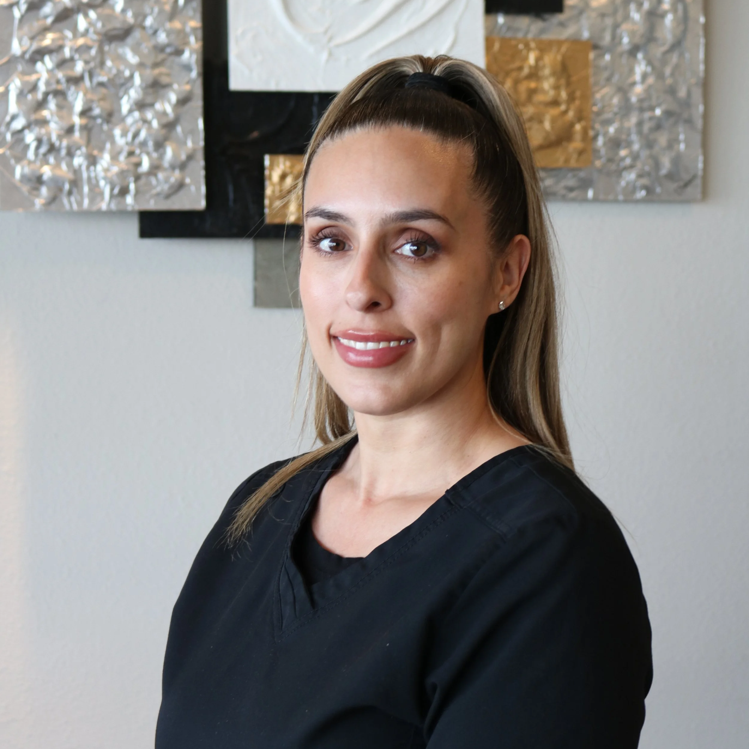 Close-up of a woman with blonde hair styled in a half-up, half-down look, smiling slightly, wearing a black top, with textured abstract art on the wall behind her.