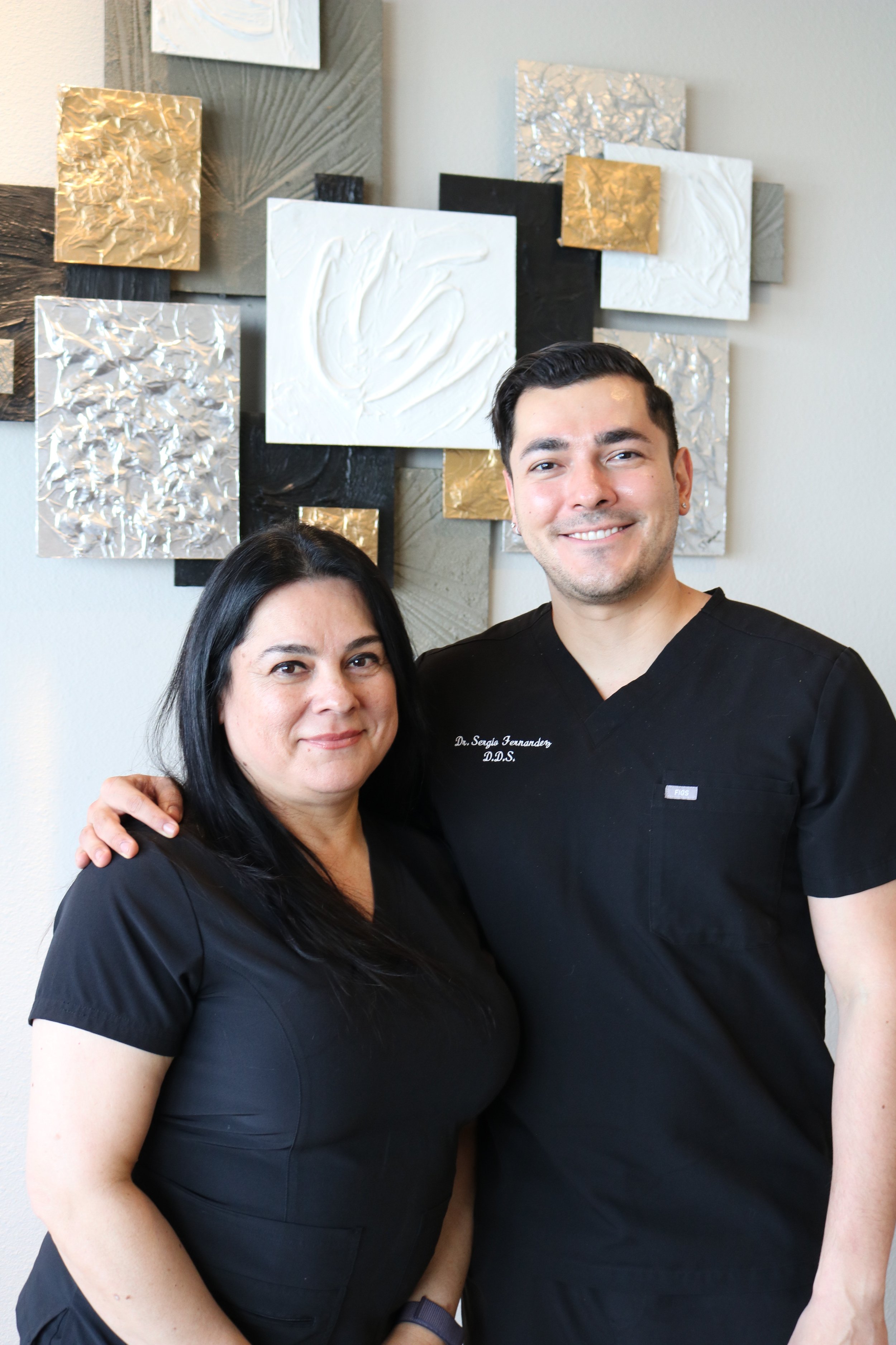 Two healthcare professionals, a woman and a man, are standing close to each other indoors, smiling at the camera with a decorative art wall in the background.