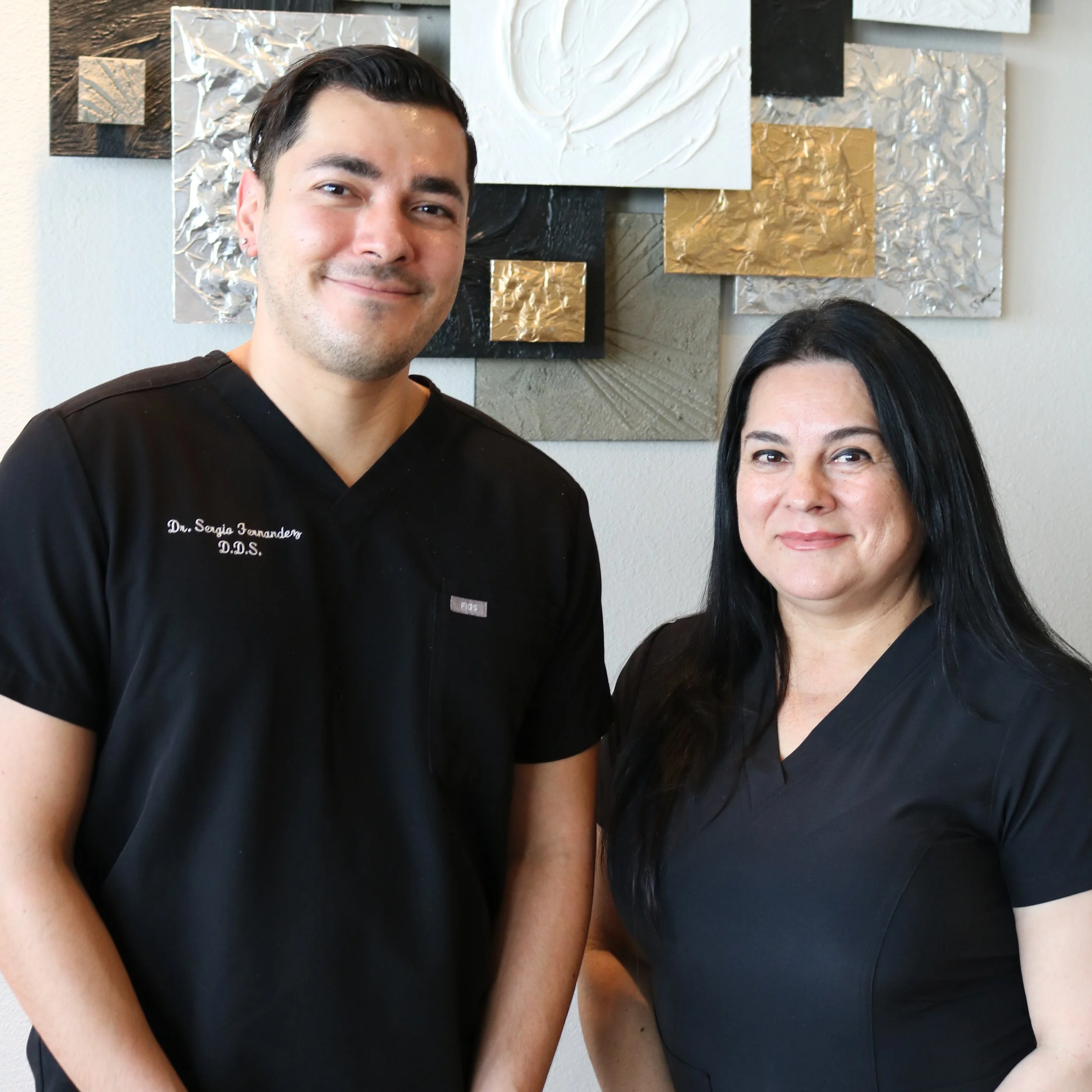 A man smiling, wearing black medical scrubs, standing next to a woman with long black hair also in black clothing, in front of an abstract wall art piece with textured square panels in black, white, gold, and silver tones.