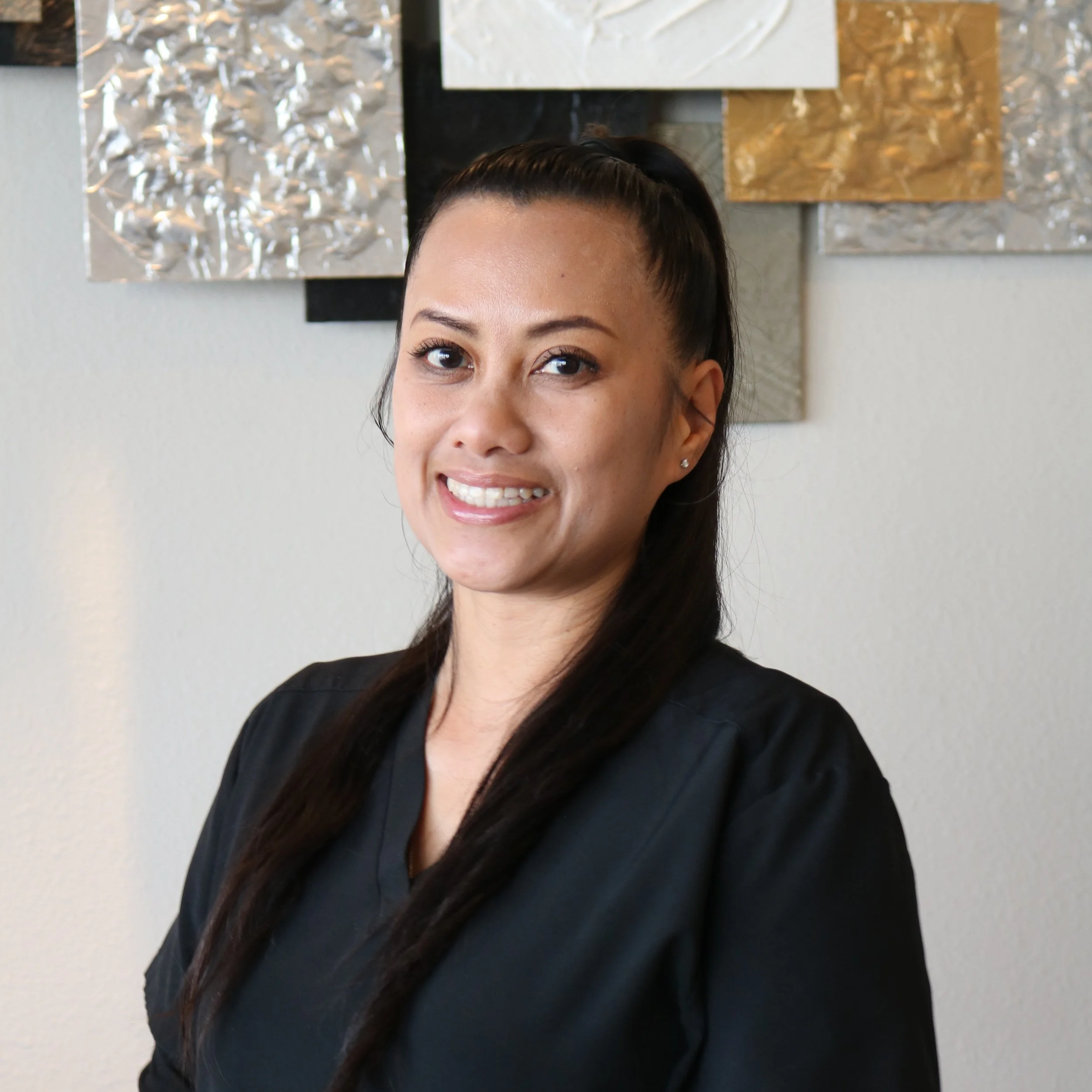 Close-up of a woman with dark hair, smiling, with a colorful textured wall art in the background.