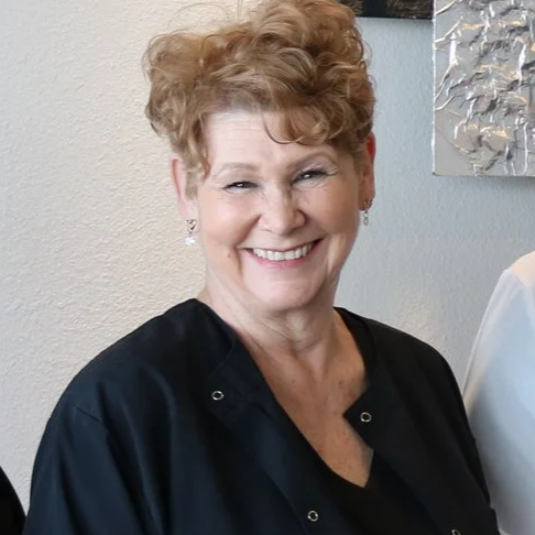 Smiling woman with short curly red hair wearing a black top, standing indoors with a light-colored wall and metallic artwork in the background.