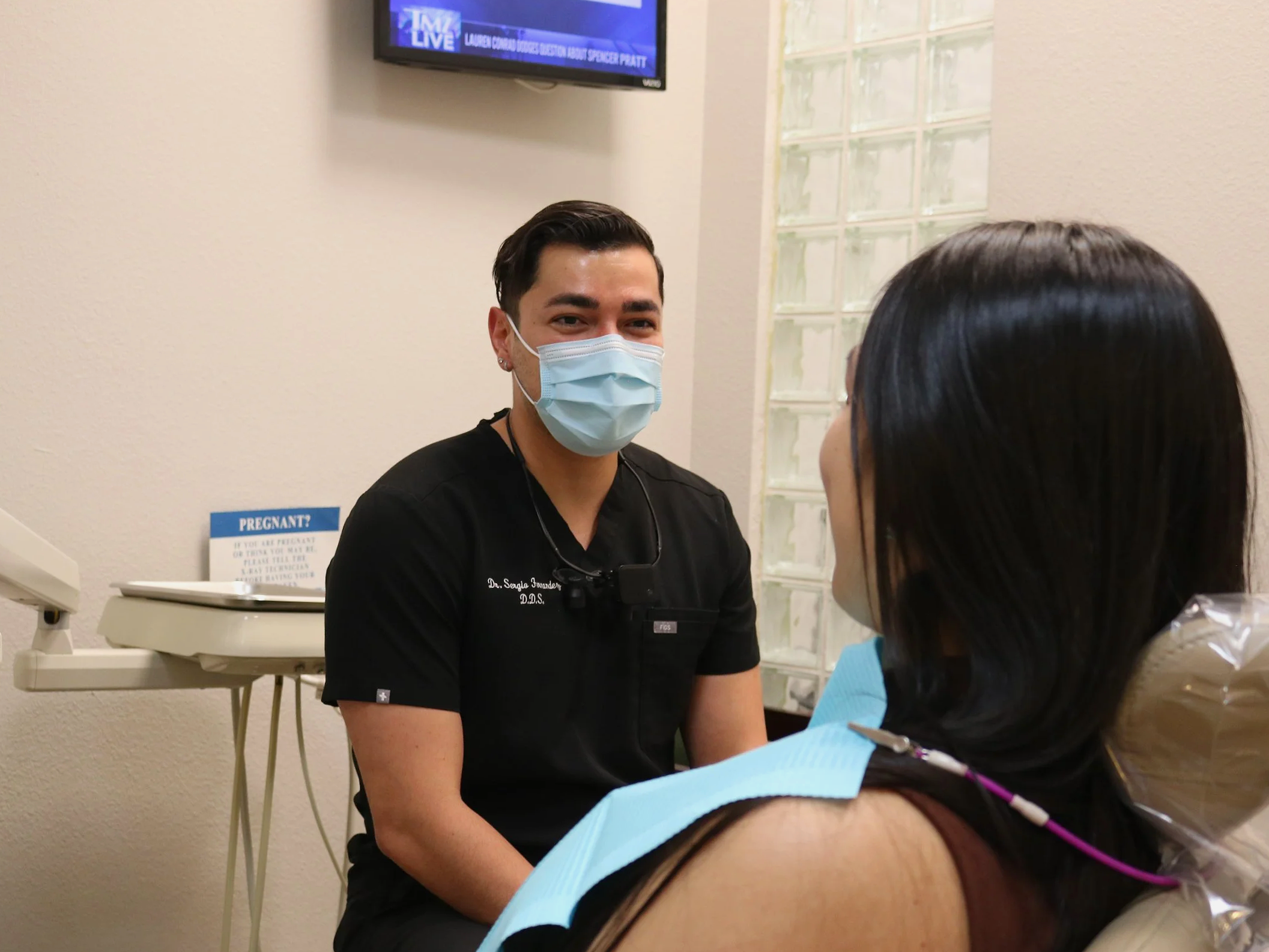 A healthcare professional wearing a face mask and black scrubs interacts with a woman in a medical setting