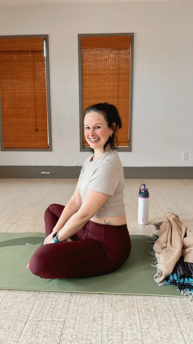 A woman with dark hair tied up, smiling and sitting on a green yoga mat indoors. She is wearing a beige t-shirt and maroon leggings. There is a water bottle and a pile of clothing or blankets beside her, and two windows with brown blinds behind her.