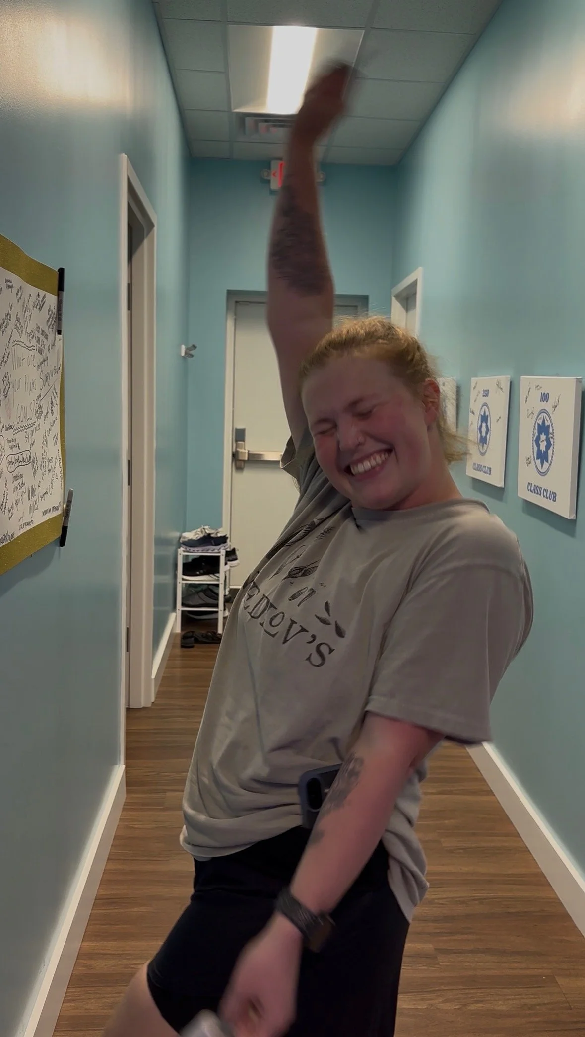 A woman in a beige t-shirt and black shorts is smiling and dancing in a hallway with light blue walls, wooden floor, and signs on the wall for a class club.