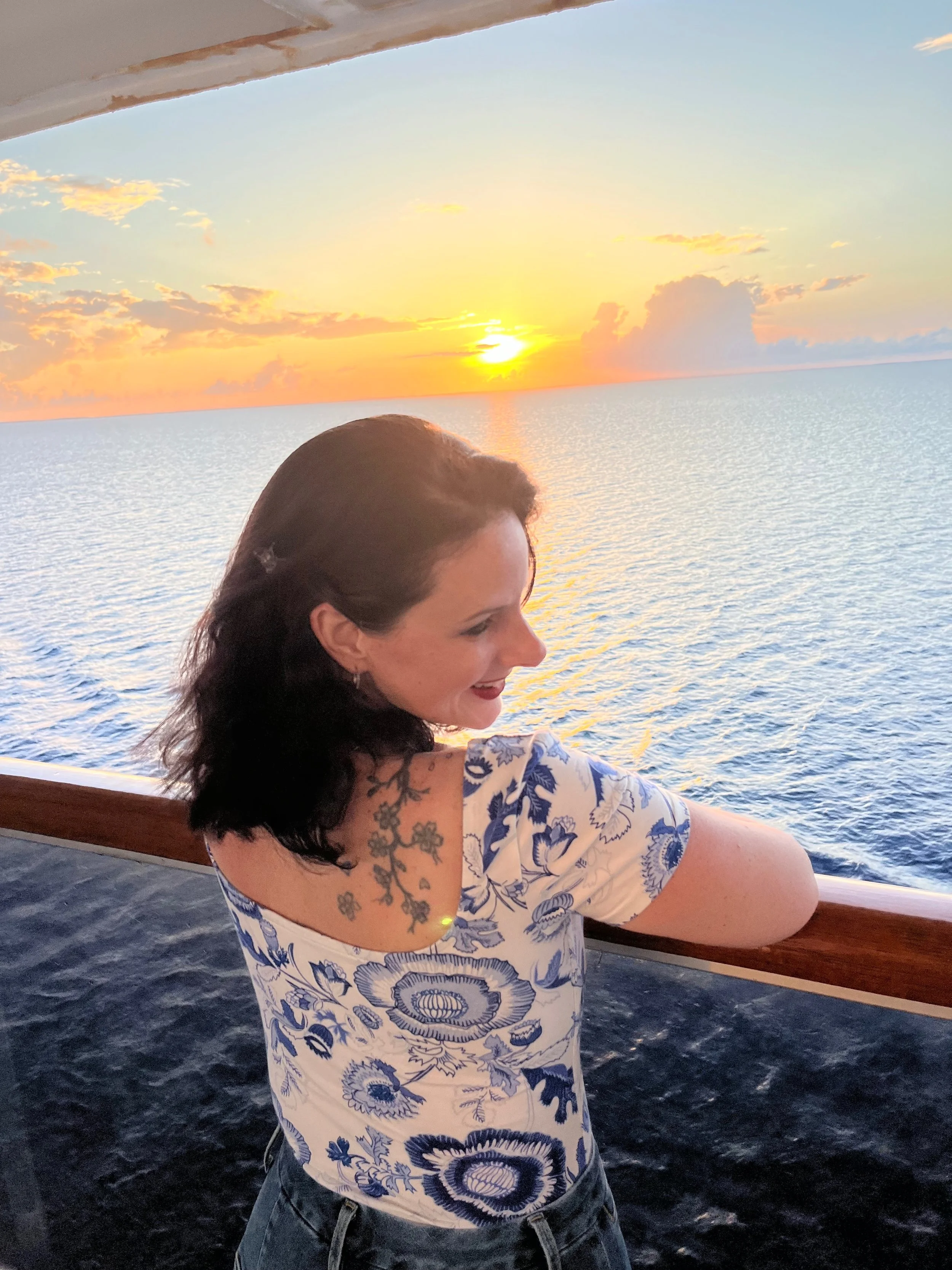 Woman with dark hair and floral tattoo on her shoulder, wearing a white and blue floral shirt, standing on a boat balcony with a sunset over the ocean in the background.