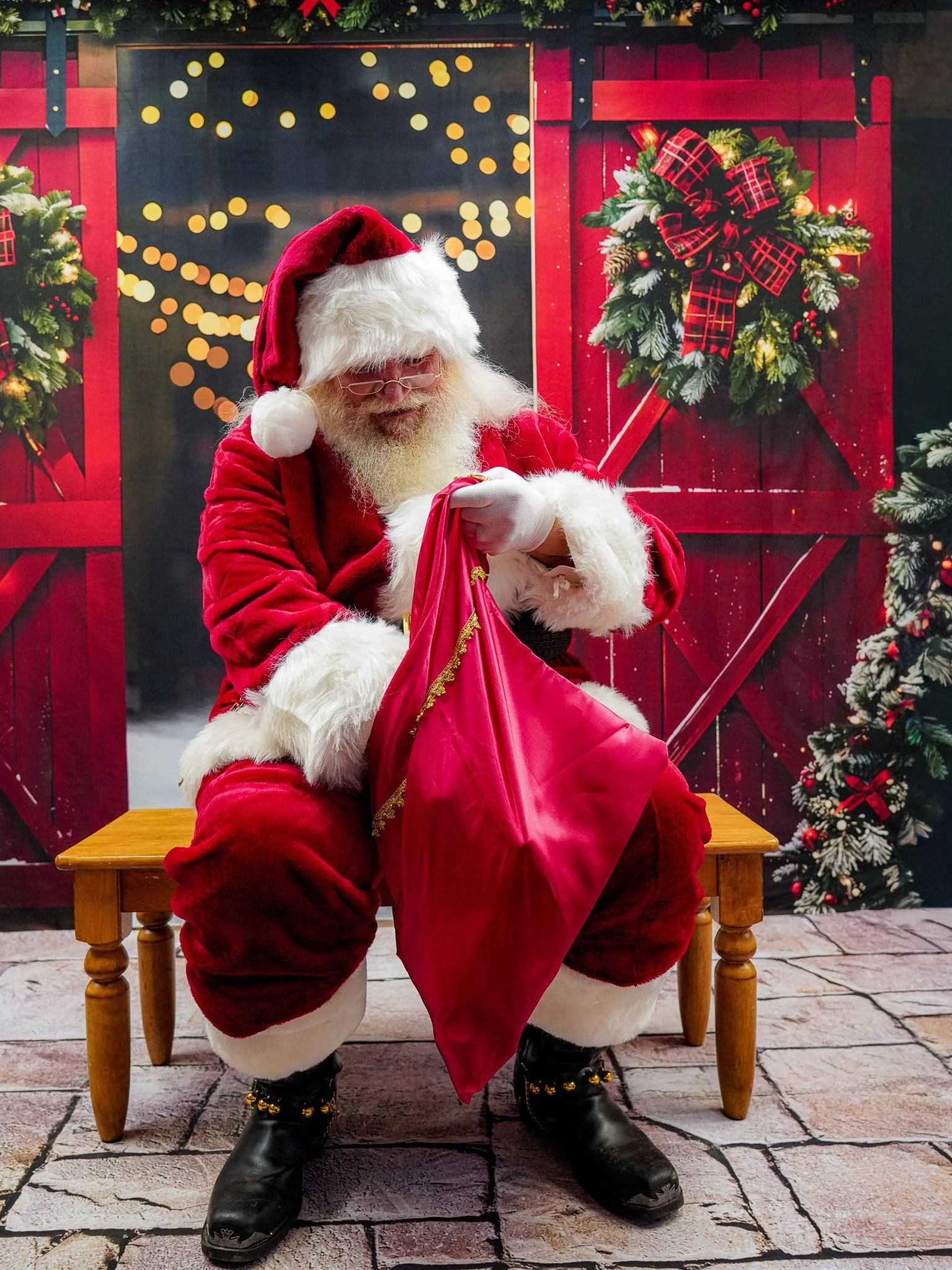 Santa Claus sitting on a wooden bench, looking into a red bag, with a festive background including Christmas wreaths, garlands, and blurred holiday lights.