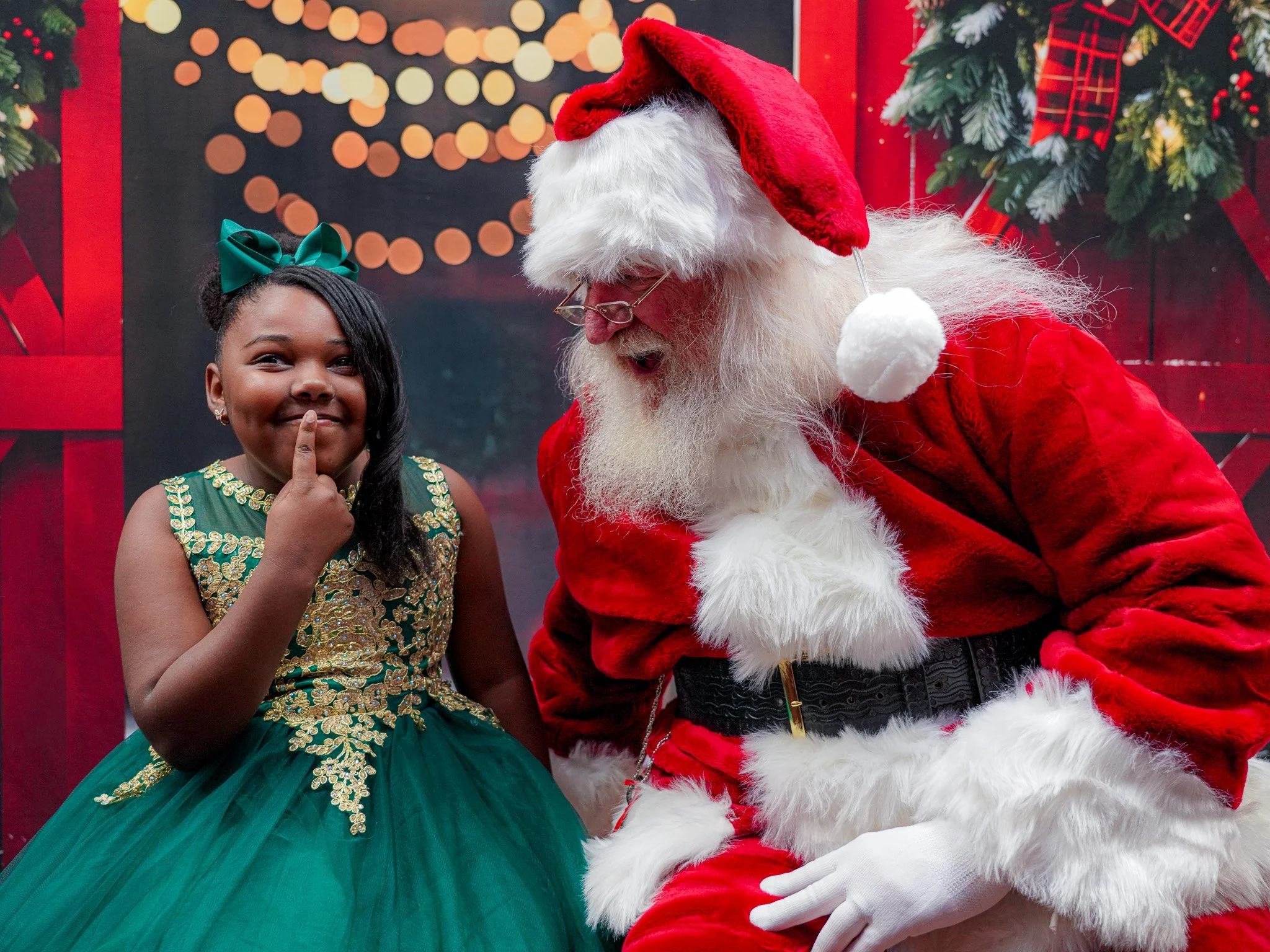 A young girl in a festive green and gold dress with a green bow in her hair is smiling while putting her finger on her lips next to Santa Claus, who is dressed in a traditional red suit with white fur trim and a Santa hat. They are in front of a Christmas-themed background with holiday decorations.